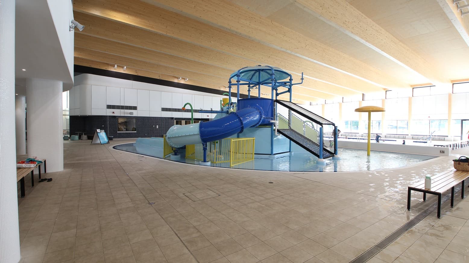 This picture shows the other side of the large aquatic centre. Kids can engage in a variety of activities while at the pool, thanks to the colorful waterpark sliding area and other kid-friendly playthings, with shades of blue and yellow. The pool has light brown lines-like ceiling that matches the multi-coloured splash park. The floor tiles also have a touch of brown. There are available long chairs on the sides of the waterpark.