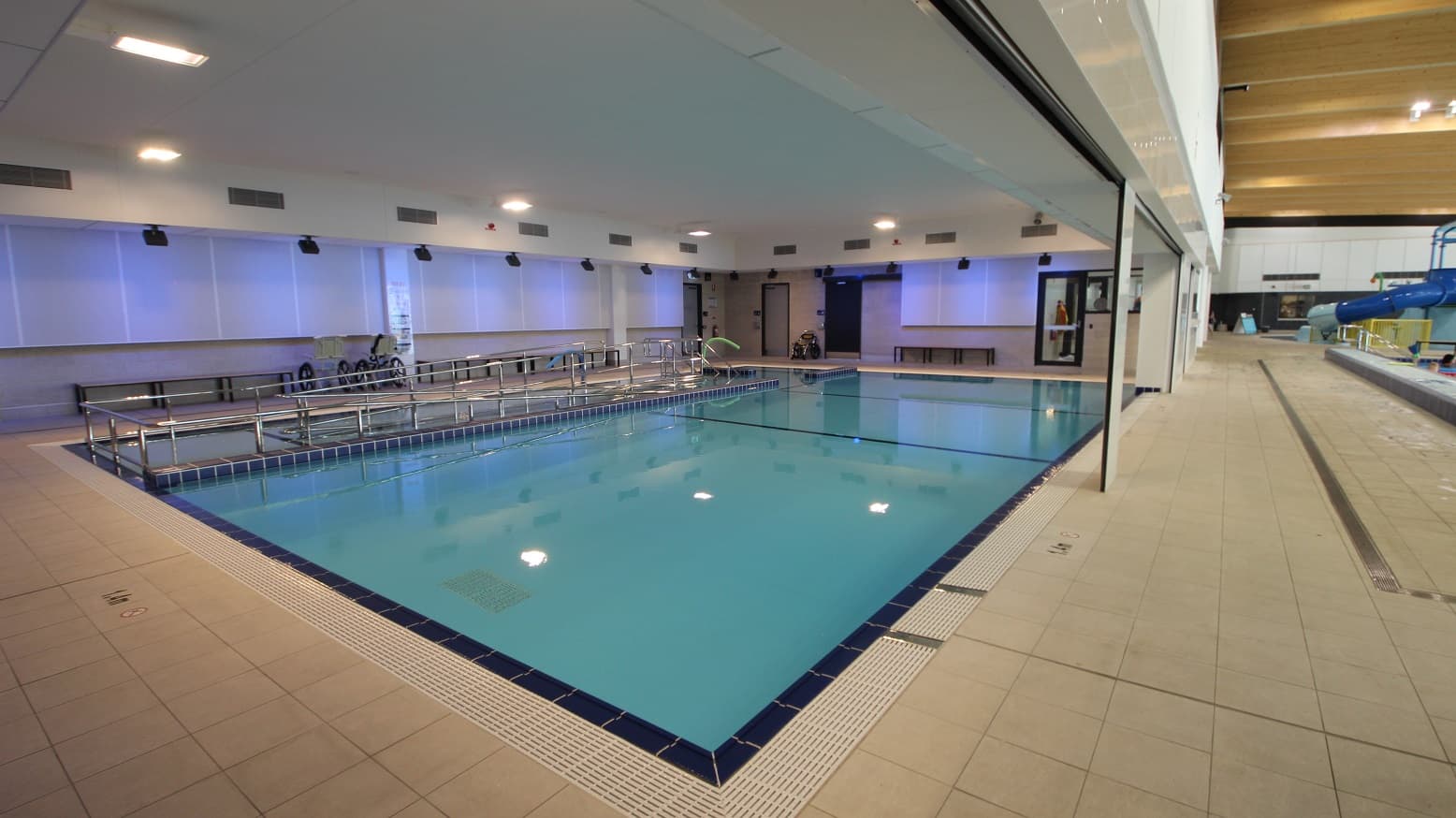 It is a large swimming pool next to the splash park that extends from one end of the area to the other with a huge white ceiling and lights. The pool has a dark blue and white frame. It has silver pool ladders and handrails that provide a safe and convenient way to enter and exit the pool. Long wooden chairs with metal legs on the side of the pool are also available.
