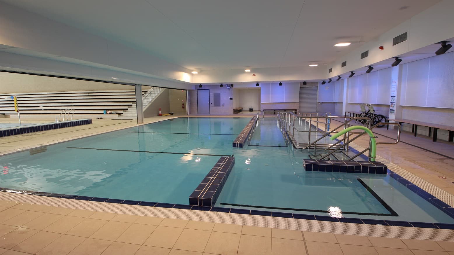 A closer look of the large swimming pool next to the splash pool park that extends from one end of the area to the other with a huge white ceiling and lights. It also has the bulkhead built in the middle of the pool that also looks like its partition. Long wooden chairs with metal legs are available by the poolside, and there are silver pool ladders and handrails that offer a secure and practical way to enter and exit the water. A standby wheelchair can be found on the side of the pool rails.