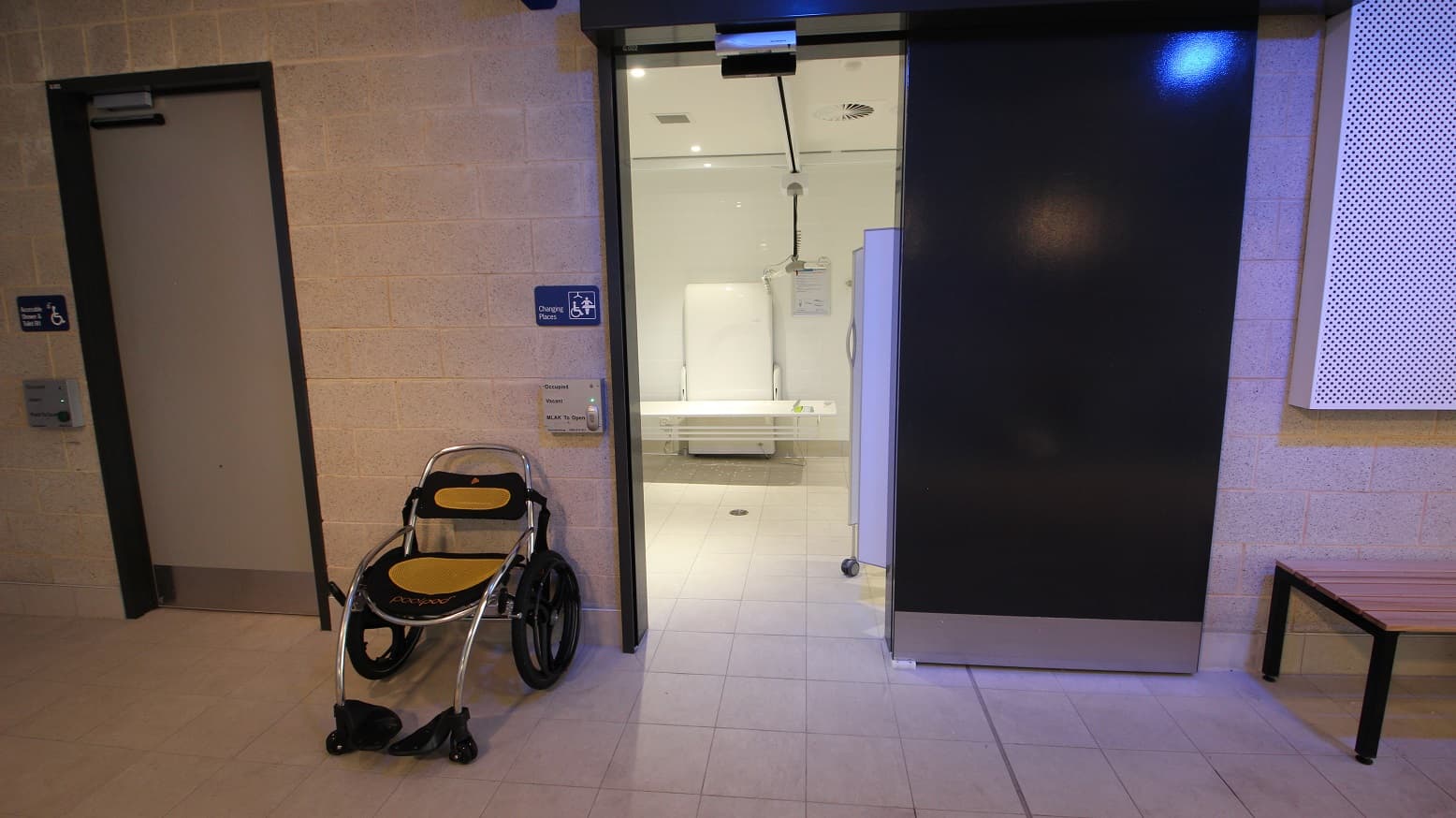 This is a glimpse of what’s inside the changing room that’s all white. What you will see while standing outside the door is the wall support and frame and the toilet bed that can be electrically raised and lowered at different heights. The room’s digital door lock mounted close to the door shows whether it is occupied or vacant and a button to open the room. A wheelchair that has rubberized seat and back support that has a combination of black and yellow color is placed outside.