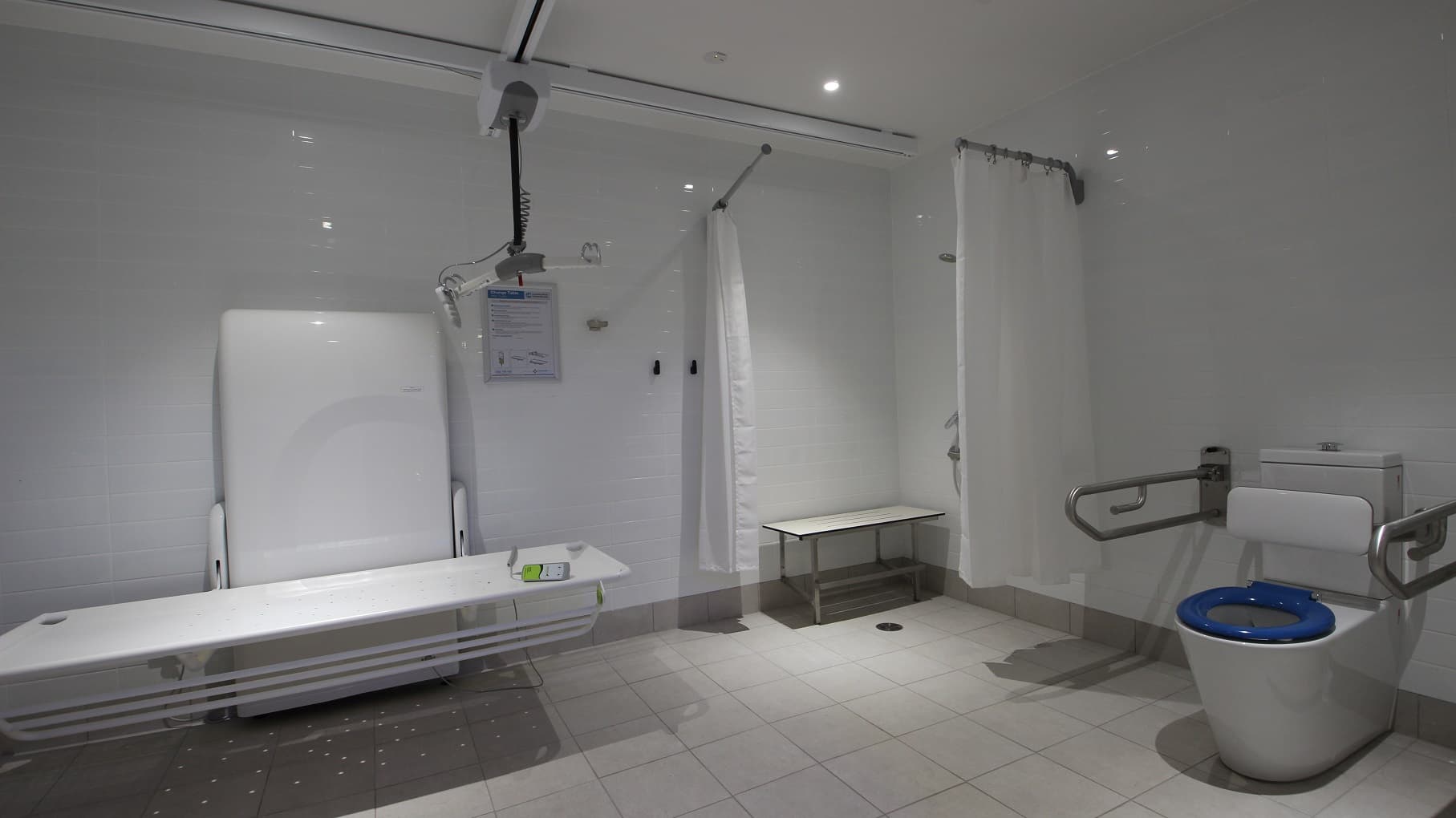 The toilet inside the Aquatic Centre section has different sections. There is a shower section that has movable white curtains. You will also find a wall support and frame to allow the toilet bed to be electrically raised and lowered at different heights. The white bed base can also be hinged up to be flat against the support as well. An instruction on how to use the white toilet bed equipment is posted on the wall to guide users.