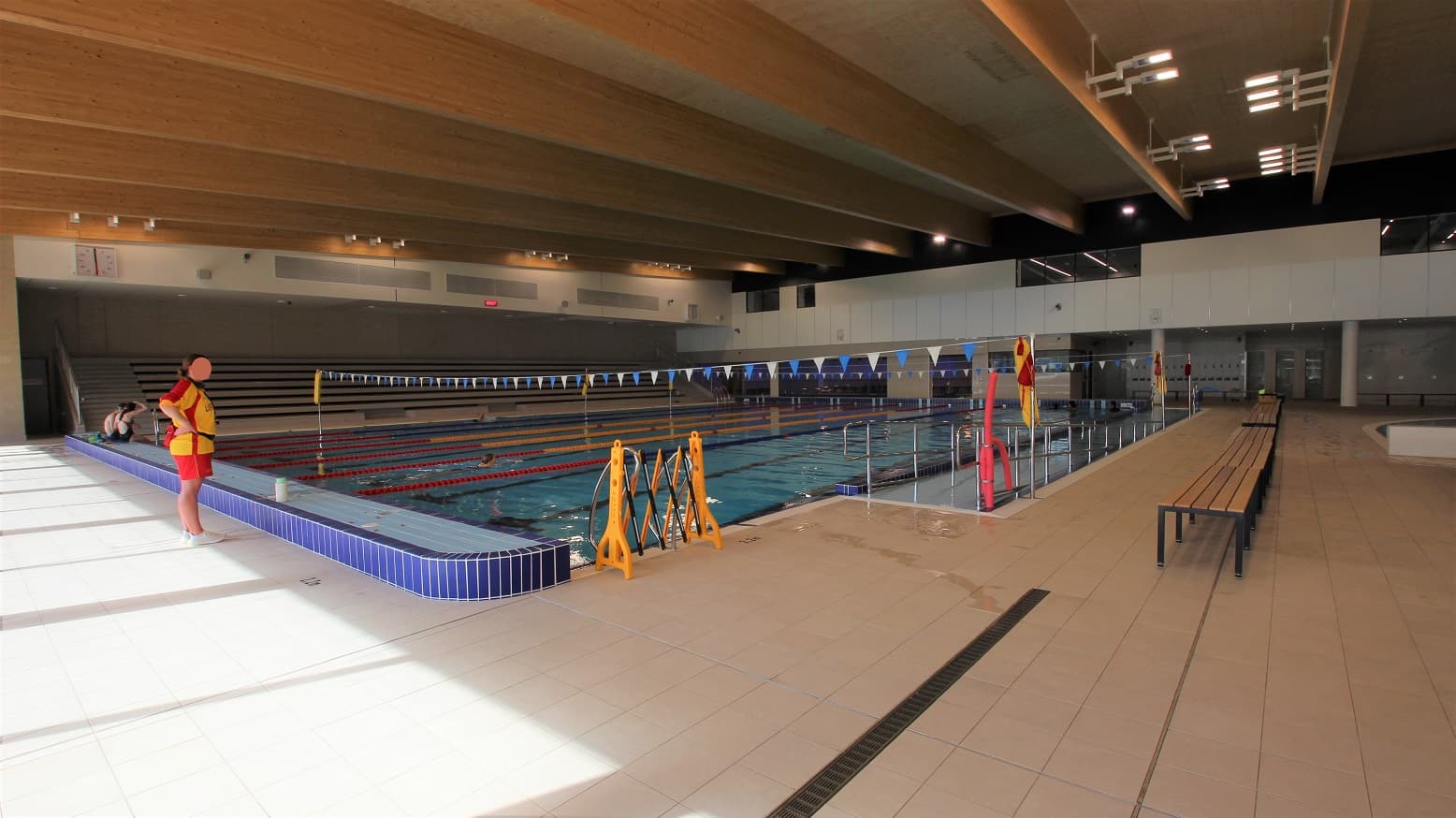 This section of the aquatic centre offers an enjoyable atmosphere and huge swimming area. A view of the competition and training pool that has three connected long wooden chairs with metal legs on the side. Banners with blue and white pennants are connected by strings hanging between the ceiling and the water surface. There are also rows of long benches for guests on the further left of the pool.