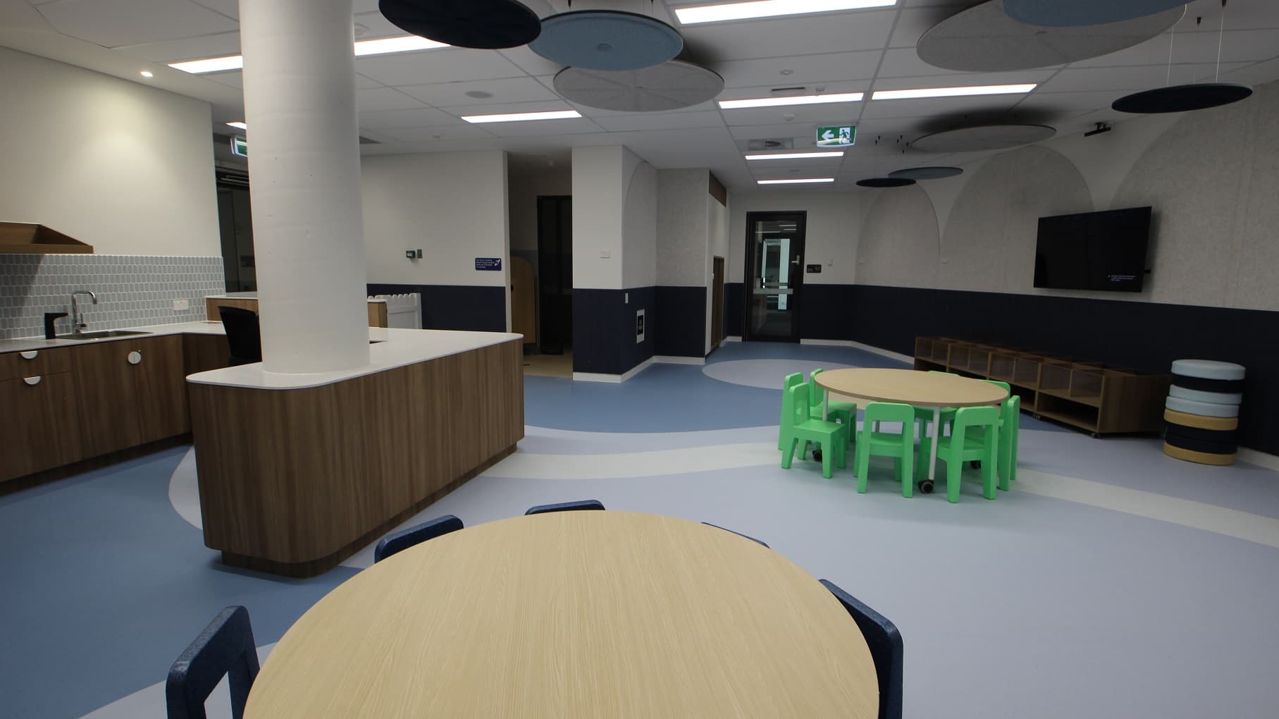 Inside the crèche are bag storage cupboards, multiple child size desks and an accessible child-size sink. The white pillar supporting the ceiling is attached to the white island counter. Close to it, are dark blue chairs tucked under the round table. At the corner, you will see a stacked round playthings with colors white, black and yellow. A wall-mounted television on the right side of the room can be seen and where a close round table and green kiddie chairs are set.