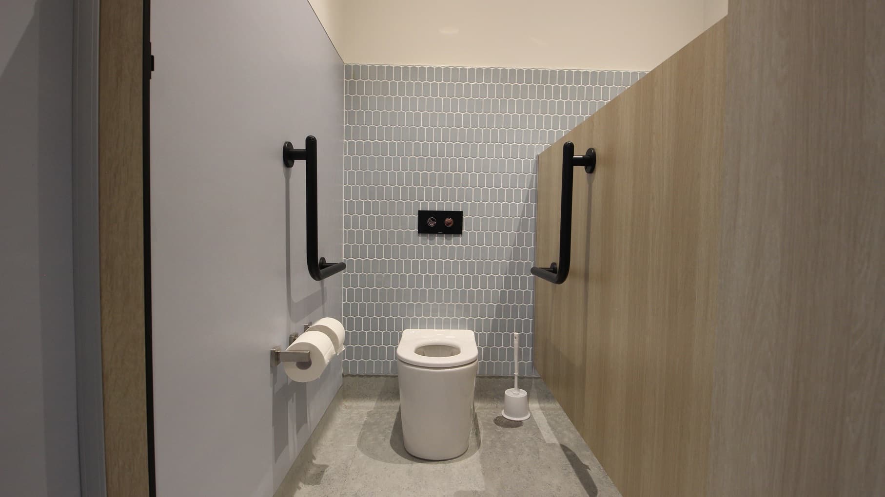 The inside view of the ambulant toilet that showcases three different wall surfaces. The right wall has a plain grey wall where the tissue holders are mounted. The back wall is designed with small grey tiles. It has a white toilet bowl with a black push button for the flush function and black handrails on both sides. The brown wall to the right is actually the partition of the two toilet units. There is also a toilet bowl cleaner placed in the corner.
