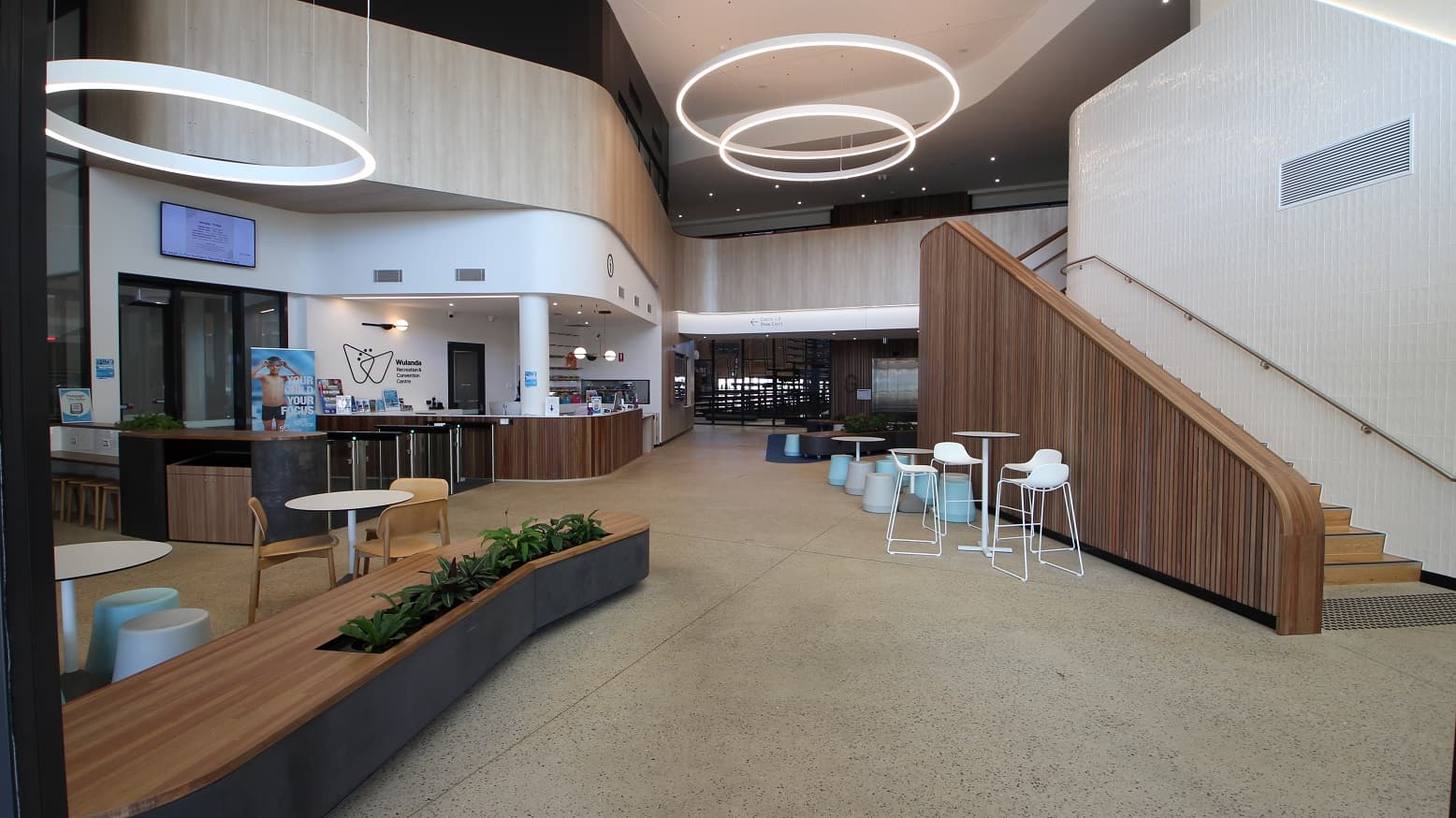 The entrance to Wulanda Recreation & Convention Centre will give you a refreshing and pleasant energy as the place is clean and has an inviting ambiance. To the right is a timber clad stairwell to access upstairs. It has large white circular fluorescent ceiling lights that serve as the focal point of the hallway. A long brown lobby bench has a built-in plant space and sets of white tables and chairs can be seen as you enter the premises.