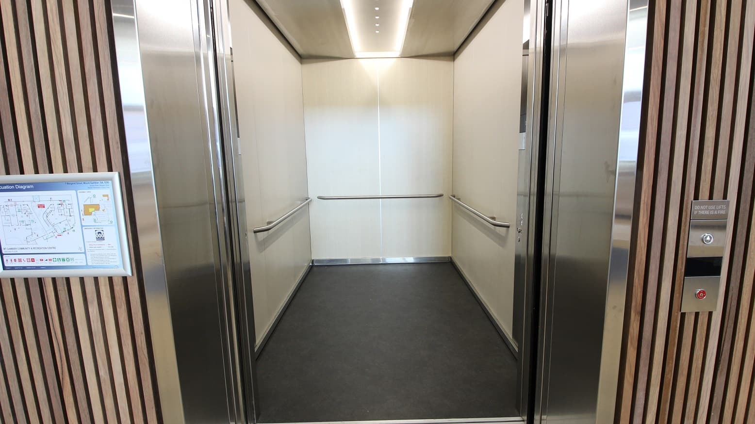 This shows what’s inside the modern elevator. The theme is minimalistic as it only has white walls and silver handrails at all sides. The floor is dark grey that complements the walls and the bright rectangular lighting inside the elevator. You will also see a closer view of the wood clad cover of the elevator’s sides, having an evacuation diagram on the left and the “UP” button with a reminder saying “Do not use lifts if there is a fire”.