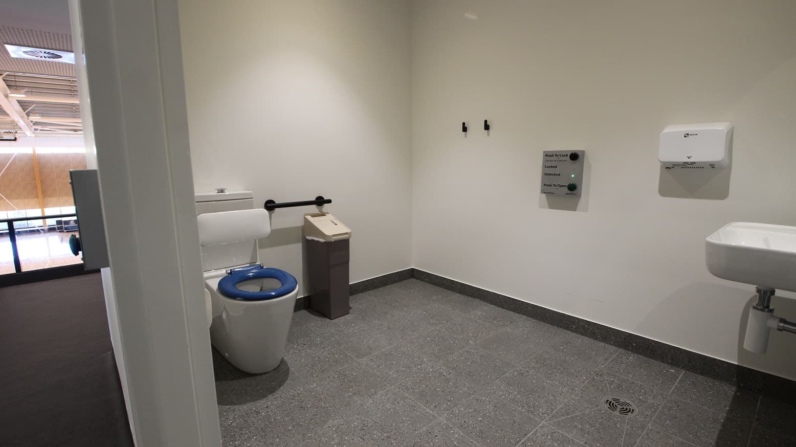 The inside view of the toilet with white walls and grey tiles. Immediate to the left is the white back to wall toilet suite that has a blue seat lid to assist guests with low vision. There is also a black trash bin with a beige cover and above it is the black grab rail. The wider wall of the toilet has two black hooks and a mounted hand dryer. In the middle part of this wall, you will find a grey digital door lock.
