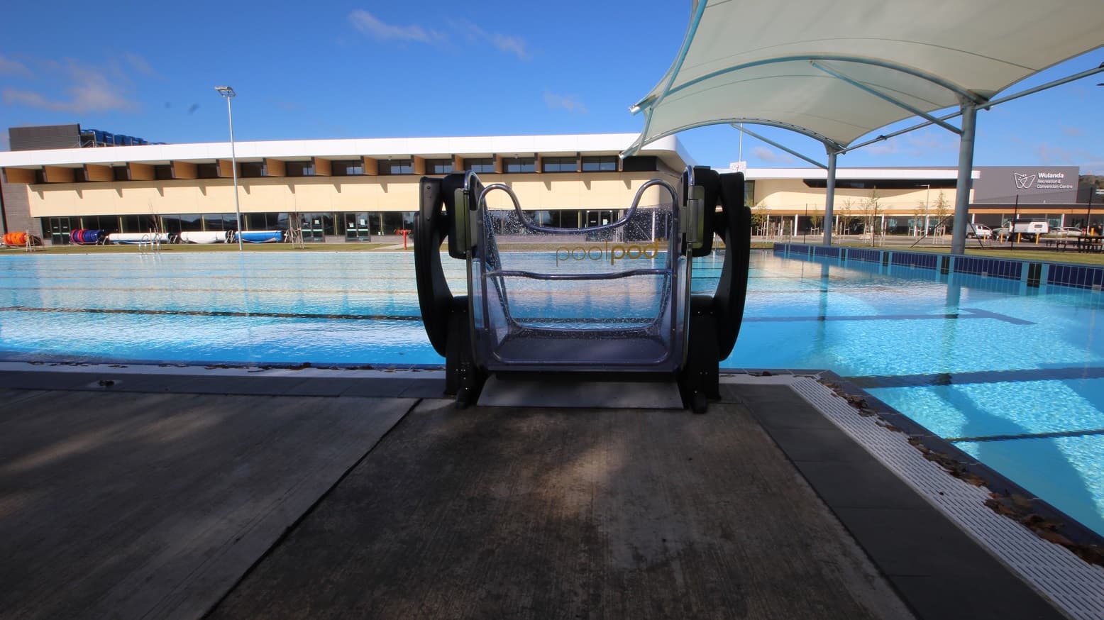 This outdoor pool showcases a pool pod that is placed on the side of the pool access stair. A white canopy is built on the right side of the pool. The lane markers are reflected on the water with long letter T-like black markings. A refreshing view from this side lets you see the indoor aquatic center and the building façade having the blue sky as its background.