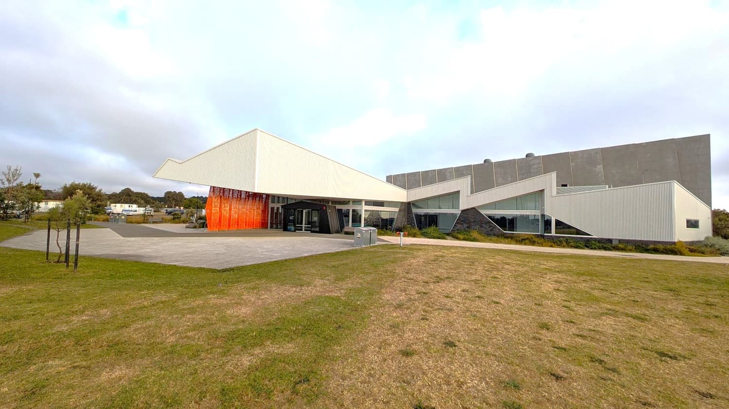 A sleek, angular white structure rises with a distinctive overhanging roof, creating a modern silhouette. Adjacent to it, an orange feature wall contrasts with the light exterior. On the right, a grey building with a textured façade features large glass windows and geometric lines, adding complexity to its form. In the foreground, a grassy area leads up to a paved space, which is backed by low trees and parked vehicles in the background, contributing to a structured yet open environment.