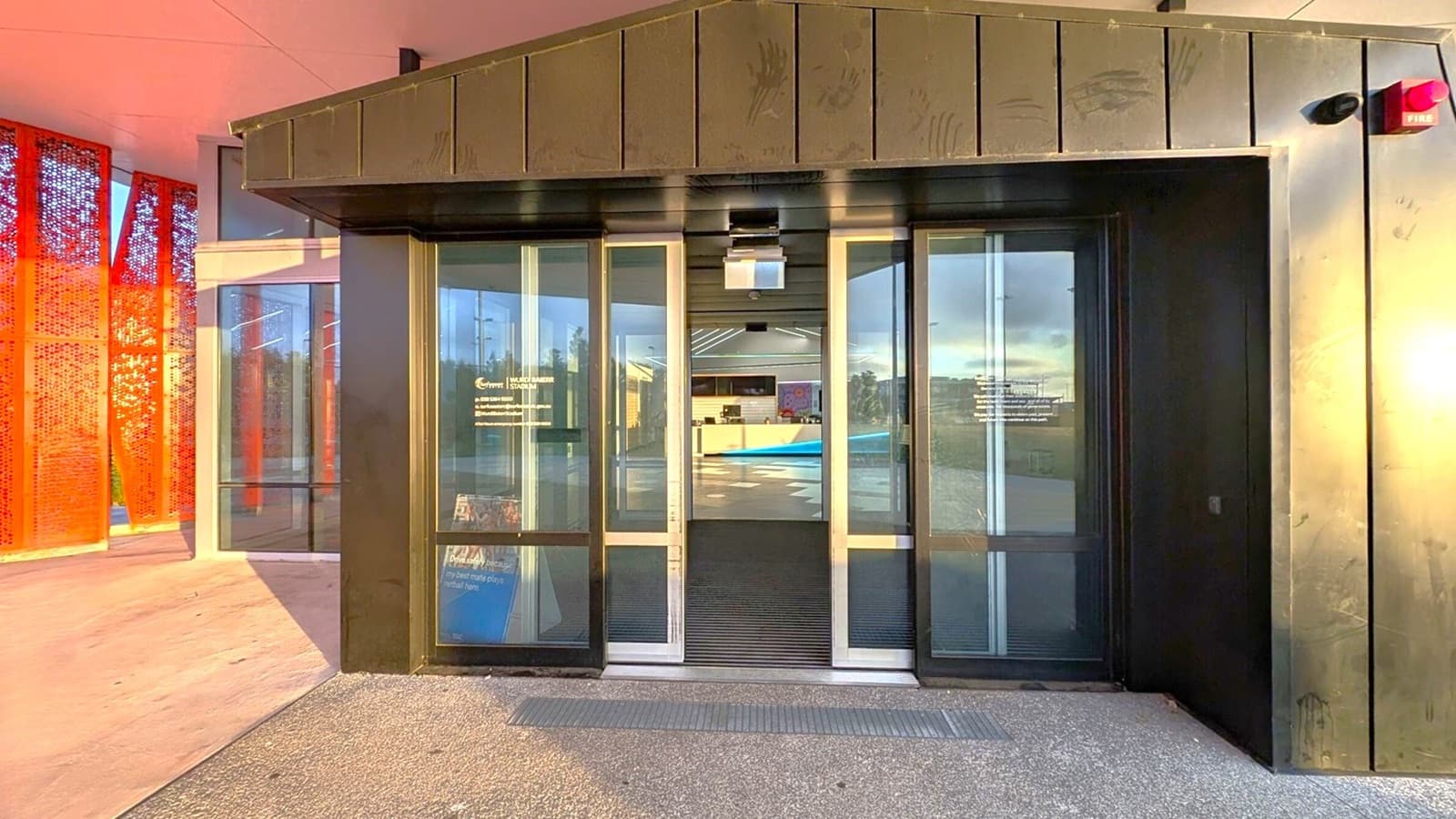 A glass entrance features sliding doors framed in black, leading into the building. To the left, a vibrant orange textured wall is visible, creating a striking contrast with the dark entrance. The floor is paved with grey material, providing a smooth surface, while the ceiling appears angular and metallic, with a slight overhang above the doorway. In the background, reflections of interior furnishings can be seen through the glass doors, indicating an open space inside.