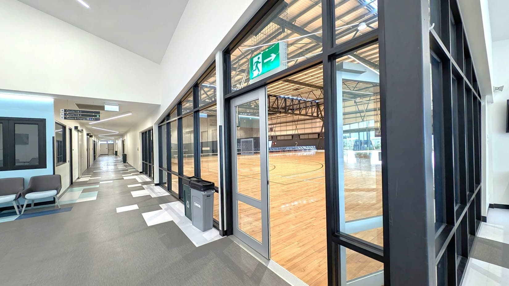 A corridor with a patterned grey and white floor and a high ceiling. On the left, there are chairs and a wall with windows. Overhead signs indicate directions to courts and facilities. On the right, large glass doors lead to a spacious indoor sports court with a wooden floor and high, metal-beamed ceiling. A green exit sign is above the door, and bins are placed nearby.