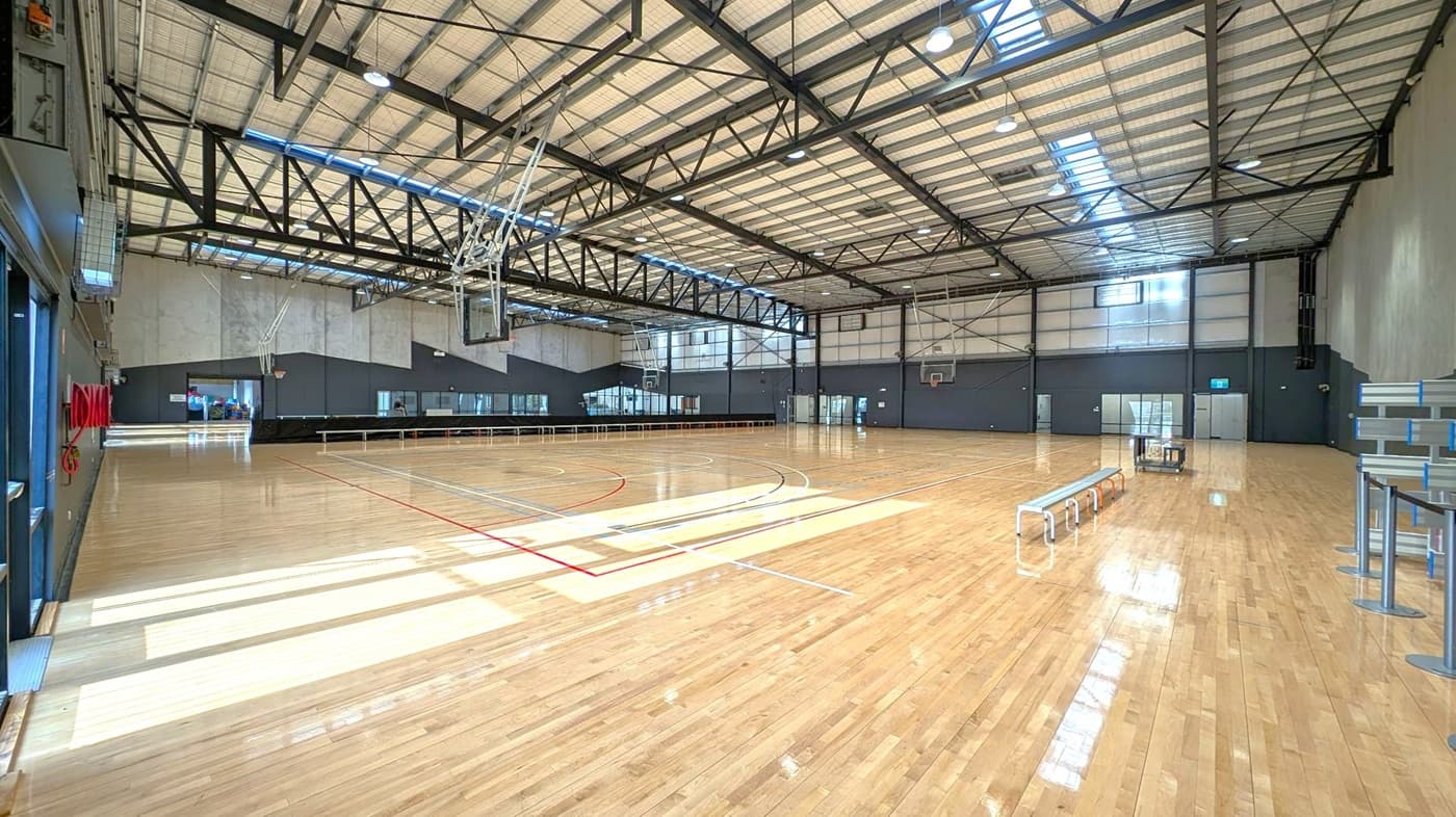 A spacious indoor basketball court with a polished wooden floor. On the left, there are wall-mounted fire hoses and a glass door. In the centre, basketball hoops hang from the ceiling, and the court is marked with various coloured lines. On the right, there are benches and a stack of chairs. The ceiling is high with exposed metal beams and skylights, allowing natural light to illuminate the space. The walls are a mix of dark and light grey.