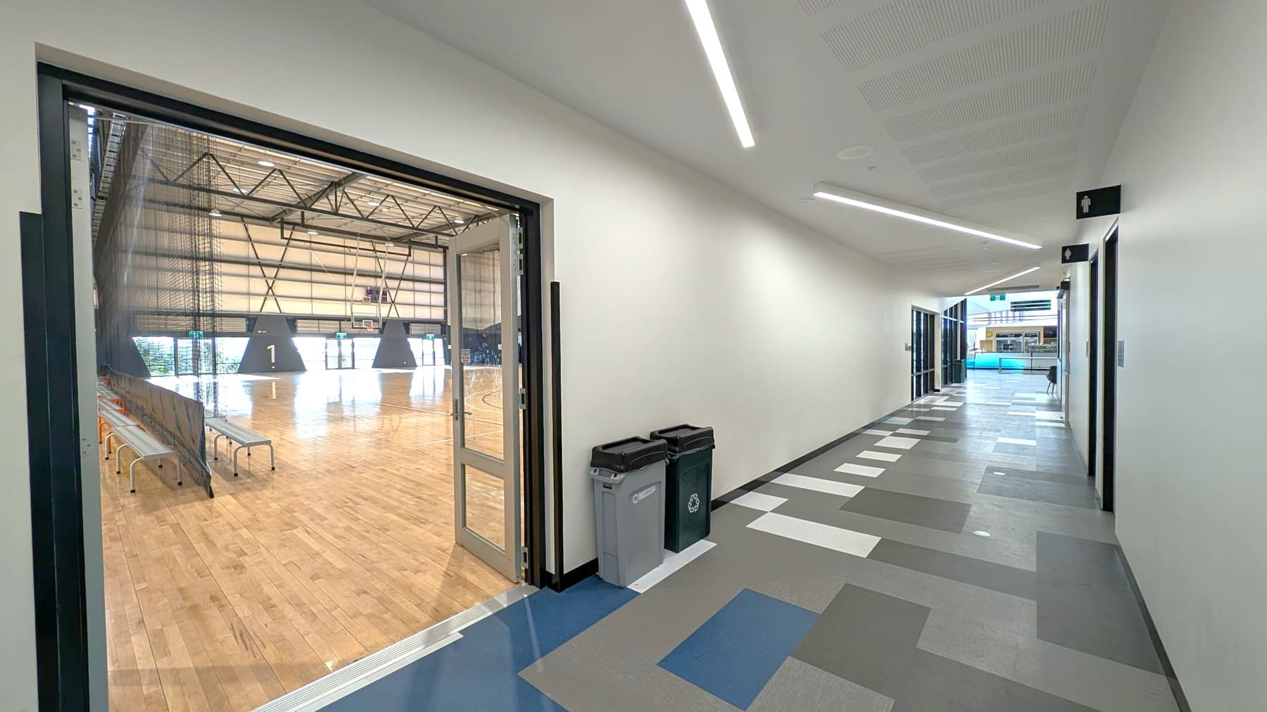 A corridor with a geometric patterned floor in shades of grey and blue. On the left, there's an open door leading to a large indoor sports hall with a wooden floor, benches, and a basketball hoop. Two bins are placed against the wall near the door. The ceiling has linear lights. On the right, there are signs for toilets, and at the end of the corridor, glass doors open to another area with visible seating.