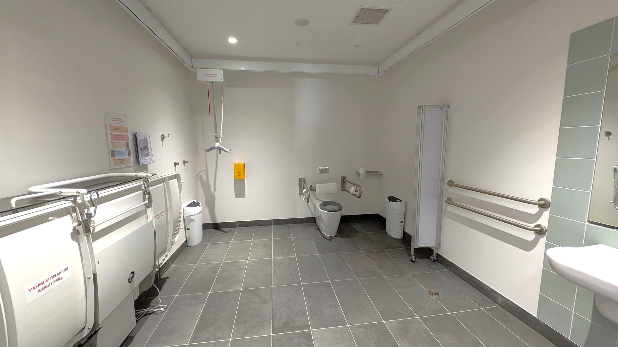 A spacious accessible bathroom with grey tiled flooring and a white ceiling. On the left, there is a foldable changing table with a sign indicating a maximum weight limit. The centre features a toilet with support rails and a wall-mounted hoist. On the right, there are two horizontal grab bars and a sink. A waste bin is positioned near the toilet, and a partial view of a mirror is visible above the sink.
