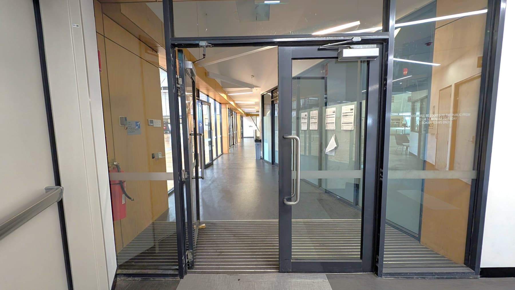 Glass double doors with metal handles open into a corridor. The floor is dark grey with a ribbed mat at the entrance. The corridor has wooden panelling on the left and glass walls on the right, displaying notices. The ceiling features linear lights. A fire extinguisher is mounted on the left wall, and a bin is visible further down the corridor.