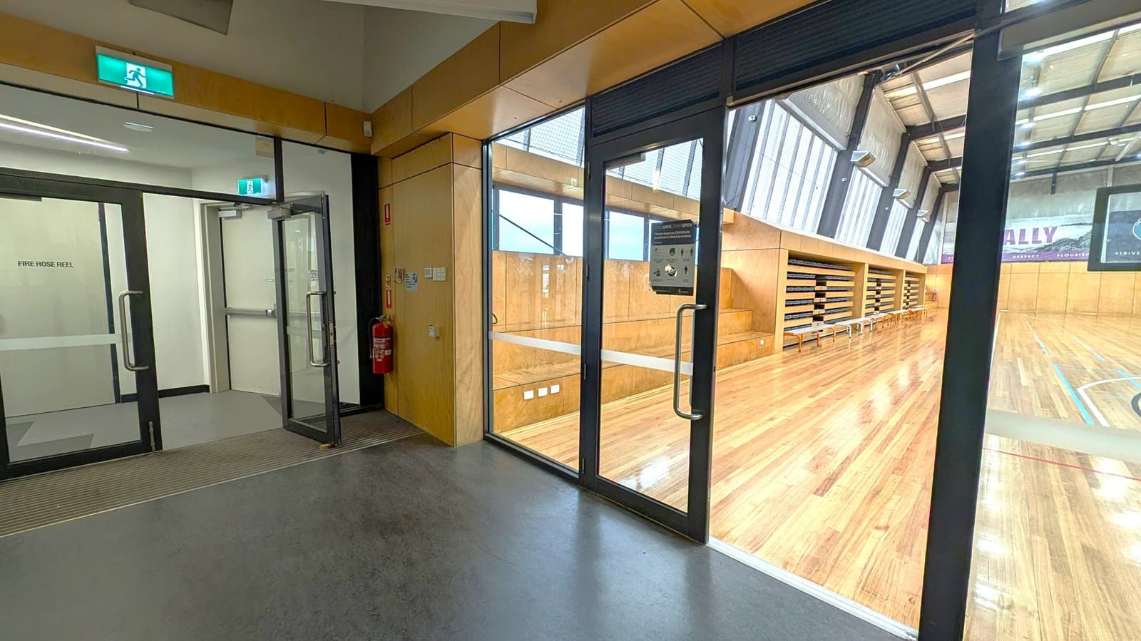 A hallway with a dark floor leads to a gymnasium. On the left, there are double glass doors with a "Fire Hose Reel" sign and an illuminated green exit sign above. On the right, large glass doors open to a wooden-floored gym with tiered seating and benches along the wall. The gym has high ceilings with exposed beams and large windows.