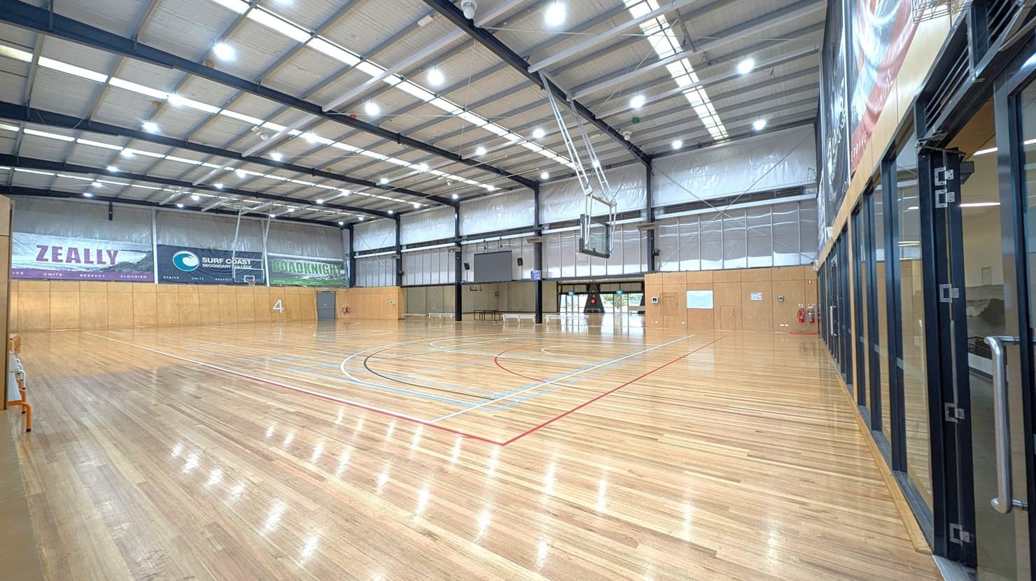 A spacious indoor sports hall with a polished wooden floor marked for basketball. On the left, there are large banners on the wall with words like "ZEALLY" and "SURF COAST SECONDARY." The ceiling is high with exposed beams and bright overhead lights. On the right, there are glass doors with metal handles. The far wall has additional banners and a basketball hoop. The hall is well-lit and empty, with no people present.