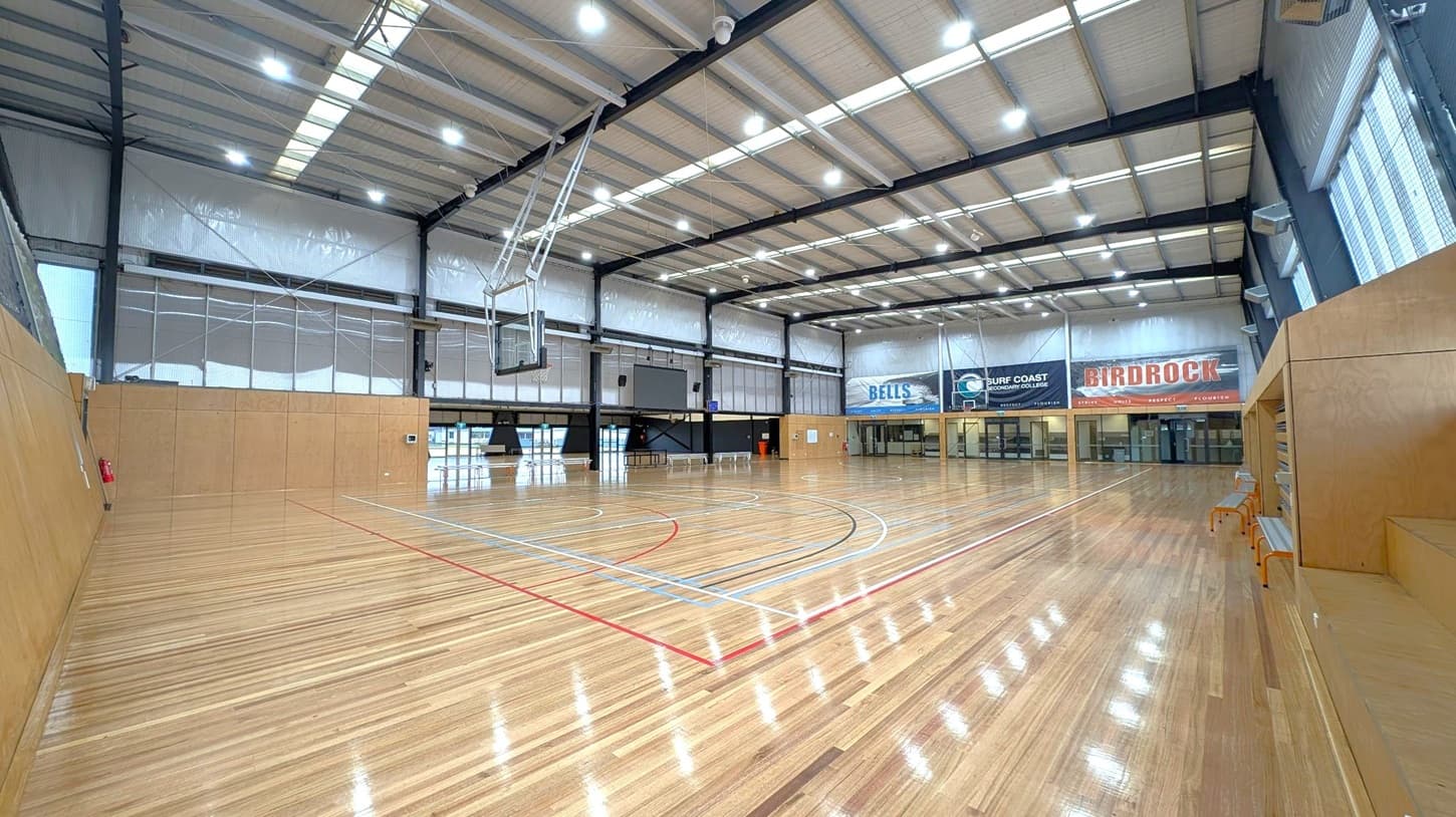 A spacious indoor basketball court with polished wooden flooring and marked lines in red, blue, and black. The ceiling is high with metal beams and bright overhead lights. On the left, there are large windows with mesh coverings. The right side features wooden benches. The far wall displays banners, including "BELLS" and "BIRDROCK," above a row of doors and windows. The basketball hoop is suspended from the ceiling.
