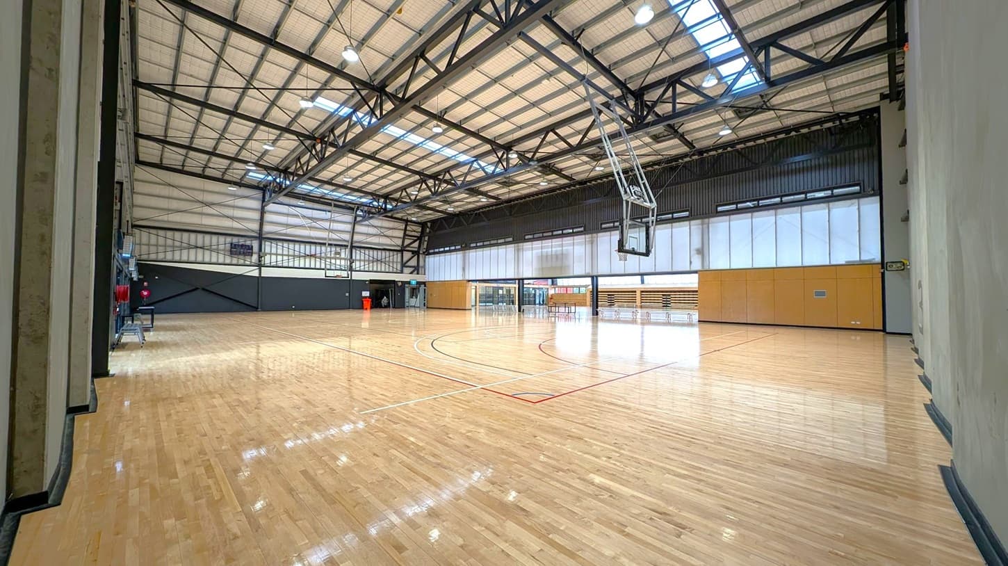A spacious indoor basketball court with a polished wooden floor. On the right, a basketball hoop is suspended from the ceiling. The left side has a dark wall with some equipment. The back wall features large windows and wooden panels, with a few chairs and a table visible. The ceiling is high with exposed beams and skylights, providing ample lighting.