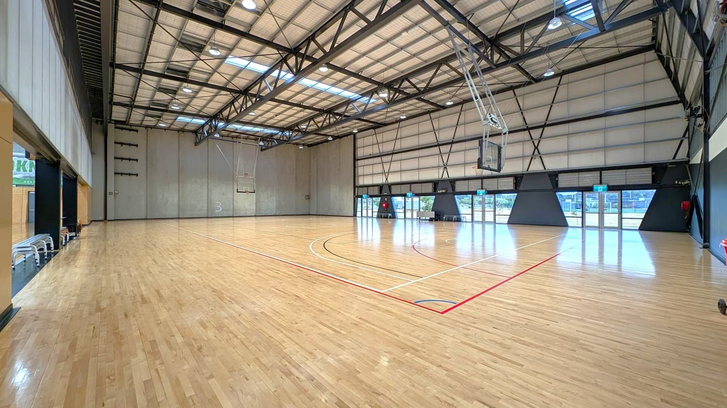 A spacious indoor basketball court with a polished wooden floor. On the left, there are retractable bleachers against a grey wall. The right side features large windows and doors with black frames, allowing natural light to enter. A basketball hoop is mounted on the right wall. The ceiling is high with exposed metal beams and several lights. The court markings are visible in red, black, and white lines.