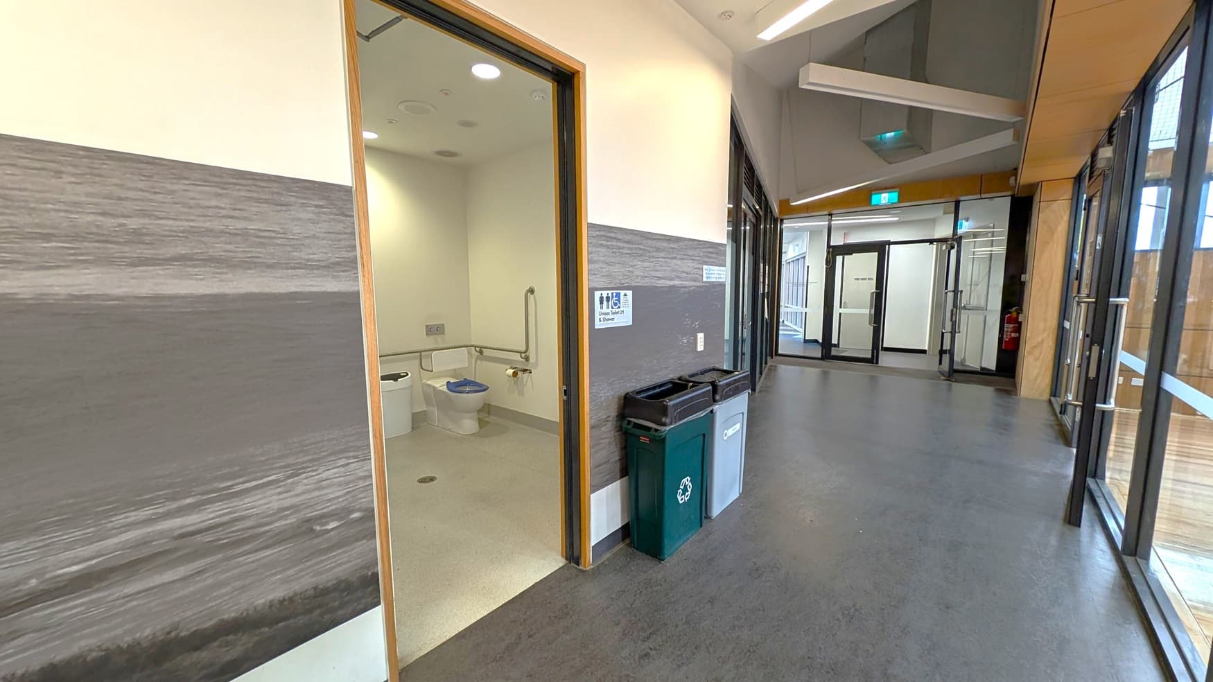 A hallway with a dark grey floor and a light-coloured ceiling. On the left, there's an open door leading to an accessible toilet with handrails. Next to the door, there are recycling and waste bins. The walls have a grey and white design. On the right, large glass windows and doors line the corridor, leading to another set of doors at the end. The space is well-lit with overhead lights.