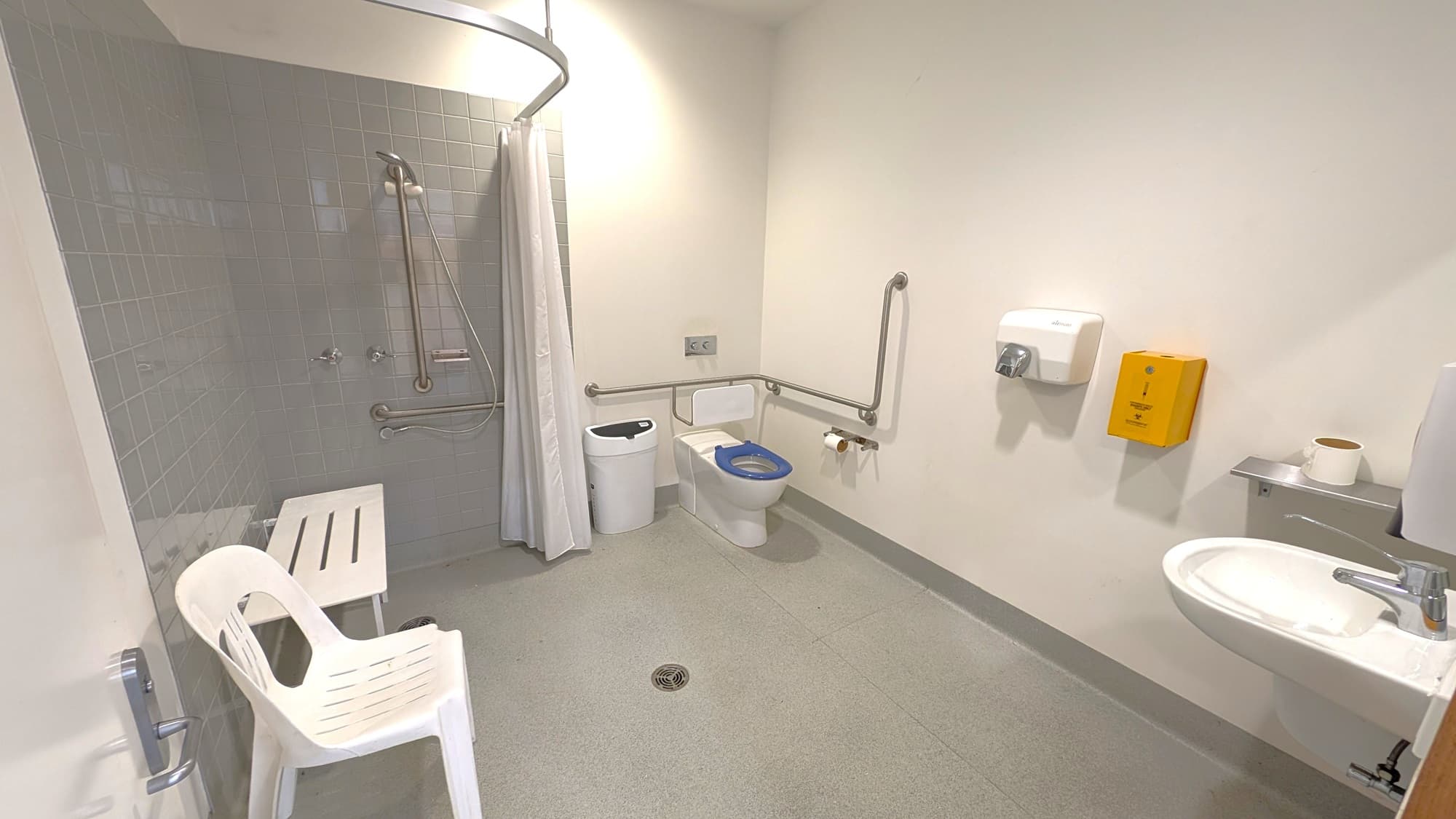 A spacious bathroom features a shower area on the left, enclosed by a white curtain with grab bars along the wall. Adjacent is a white plastic chair and a small white bench. On the right, there’s a standard toilet with grab bars, a toilet roll holder, and a bin nearby. Above the toilet, there are controls mounted on the wall. To the right, a soap dispenser and a yellow emergency kit are visible. The floor is grey and smooth, while the ceiling appears plain and white.