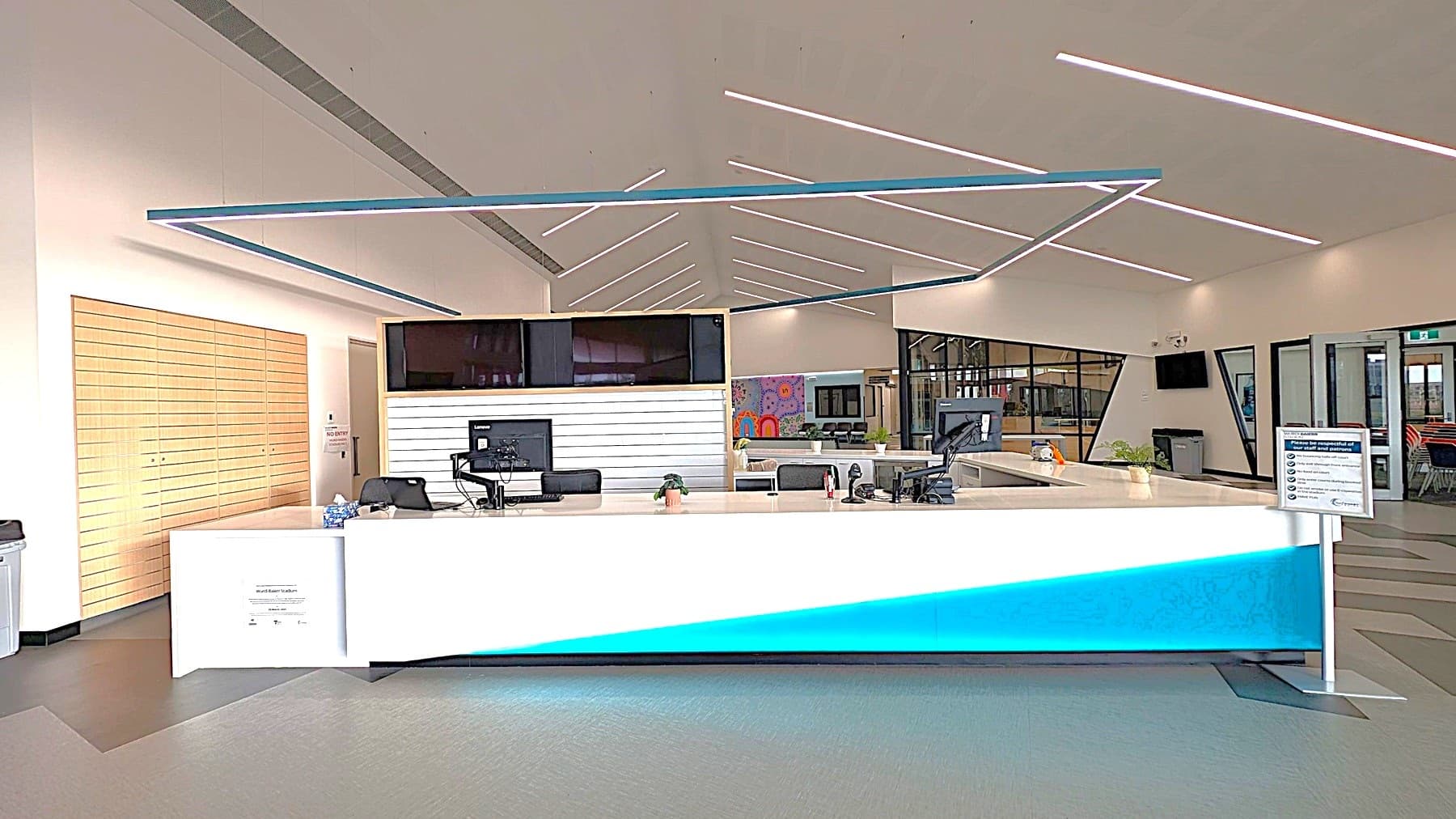 A modern reception area with a sleek white desk featuring a blue accent light at the base. On the desk are computers, a small plant, and office supplies. Behind the desk, there's a wall with horizontal wooden panels on the left and a glass partition on the right. The ceiling has geometric lighting patterns, and the floor is a mix of grey and black tiles. In the background, there are seating areas and a colourful mural.