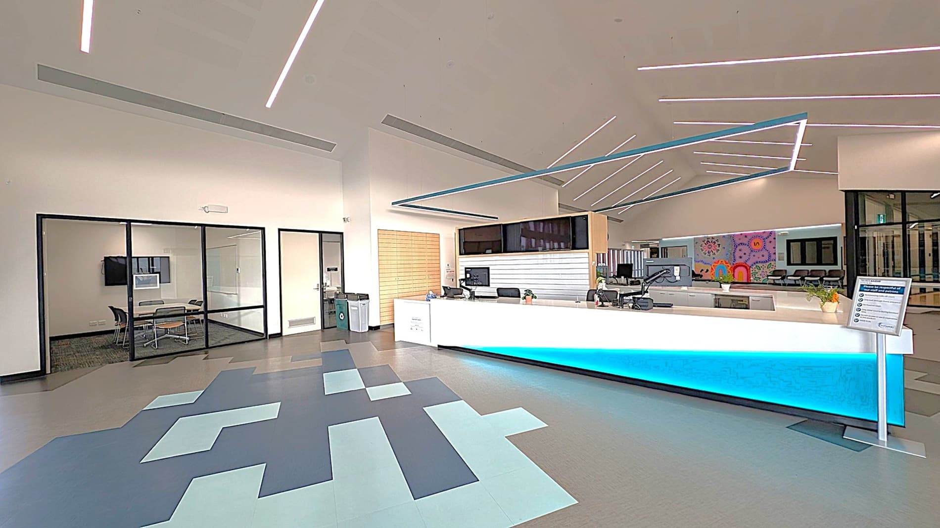 A modern reception area with a sleek, white desk illuminated by blue lighting underneath. On the right, there are computer monitors and office supplies. To the left, a glass-walled meeting room with a table and chairs. The floor features a geometric pattern in shades of grey and teal. The ceiling has linear LED lights arranged in a dynamic pattern. In the background, a colourful mural is visible on the wall.