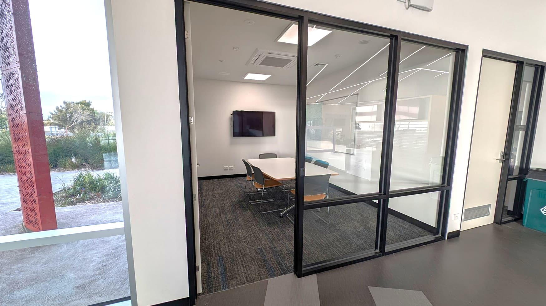 A small meeting room with glass walls and a black frame. Inside, a rectangular table is surrounded by several chairs. A flat-screen TV is mounted on the white wall. The floor is carpeted in dark grey. Outside the room, the floor is a darker shade with a geometric pattern. To the left, there is a large window showing a view of greenery and a perforated red metal structure. The ceiling has recessed lighting and air conditioning vents.