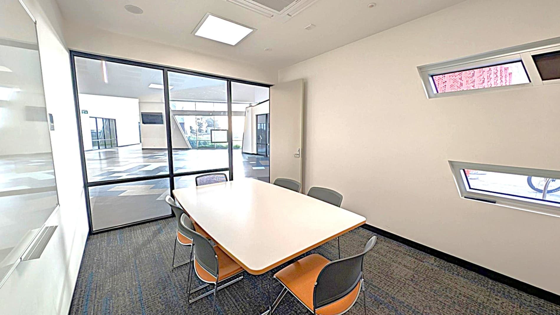 A small meeting room with a rectangular table surrounded by six chairs. The room has light green walls and a dark carpeted floor. On the right wall, there are two narrow, horizontal windows. The left side features a glass wall with a door, providing a view into a larger, open area with a patterned floor. The ceiling has recessed lighting.