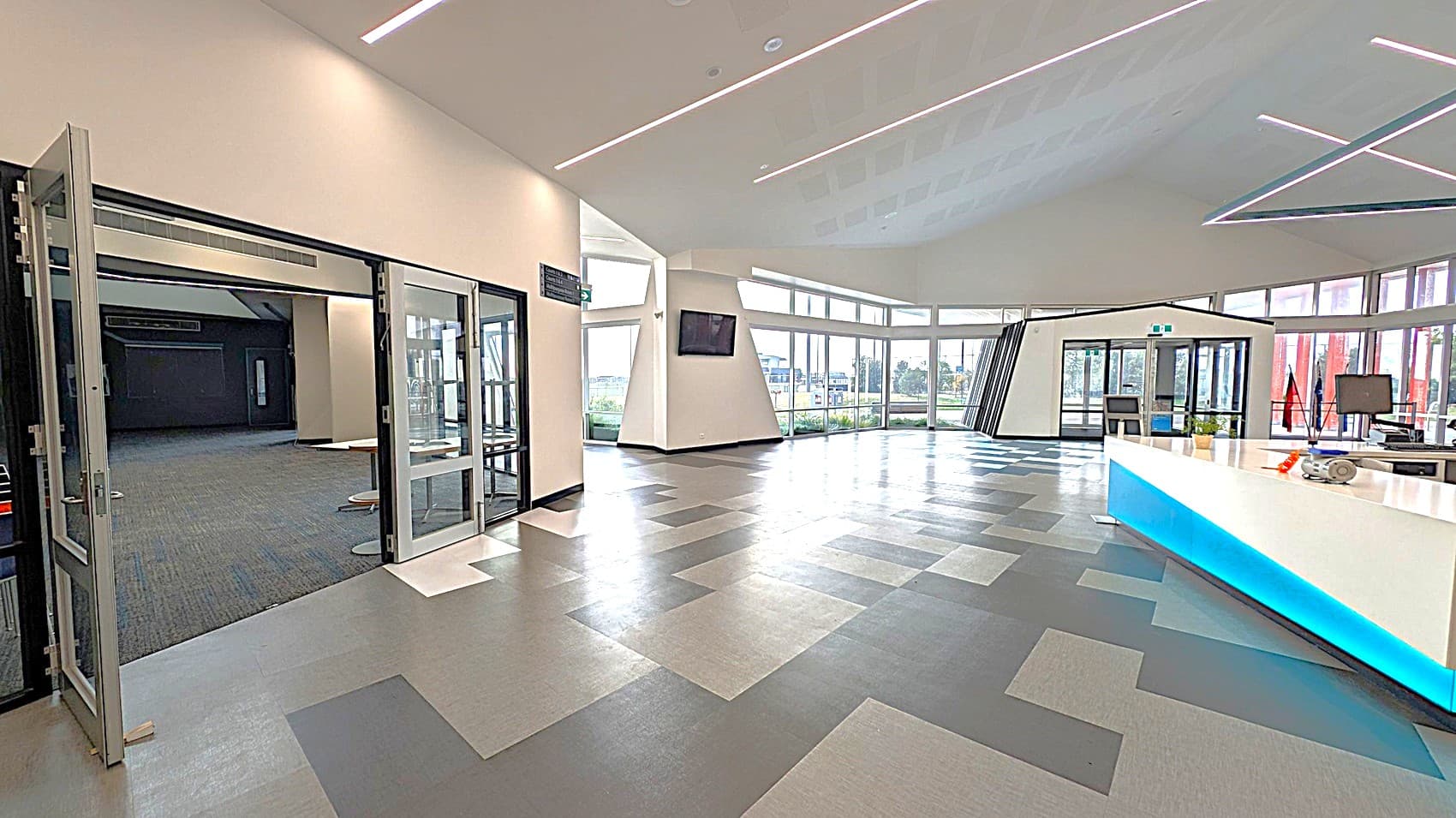 A spacious, modern interior with a geometric patterned floor in shades of grey. On the left, there are open glass doors leading to a carpeted area. In the centre, large windows provide a view of the outdoors, with a triangular column and a wall-mounted screen. On the right, a white reception desk with a blue accent light, and a small plant on top. The ceiling features linear LED lights.
