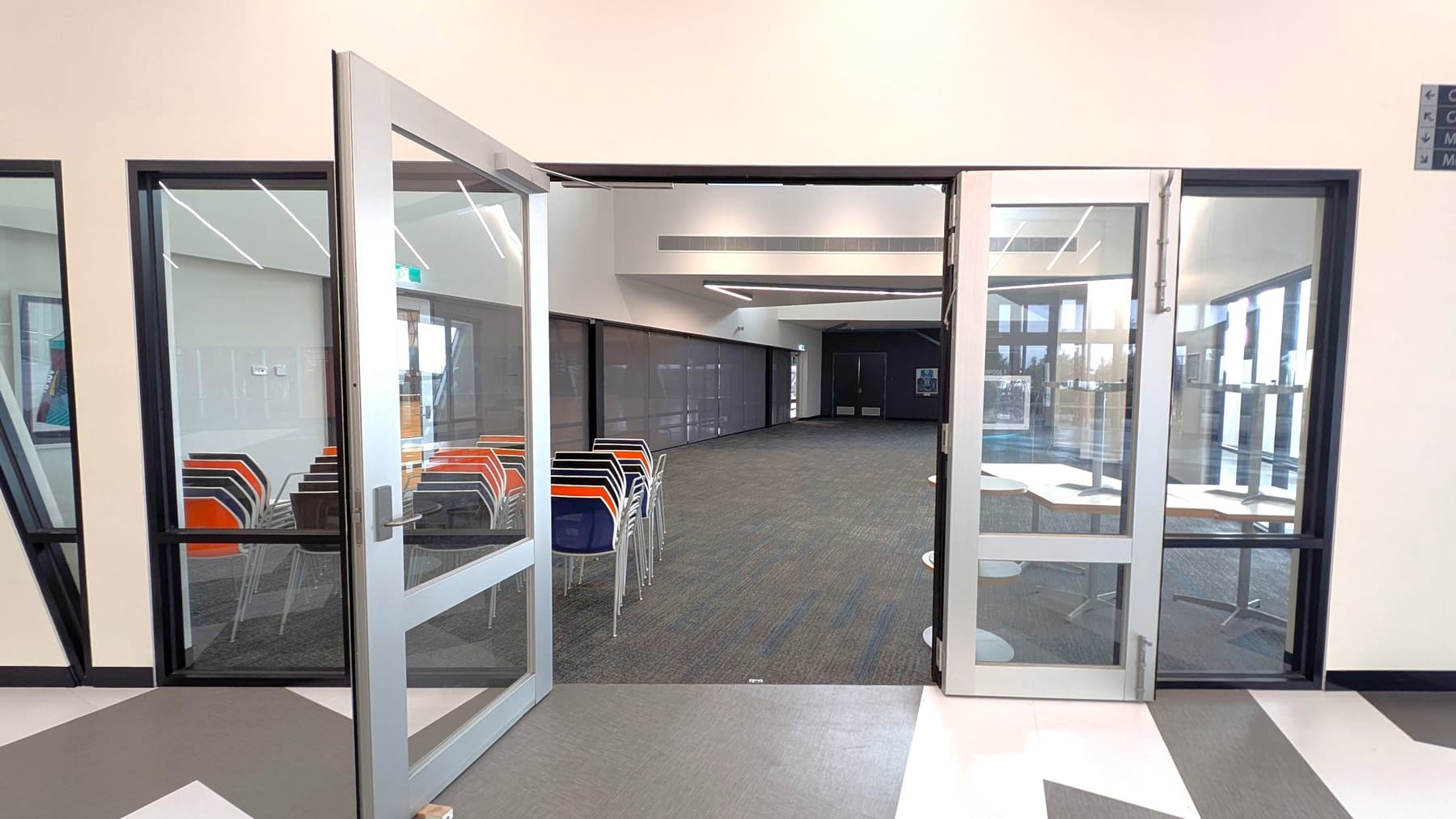 A modern interior space with glass double doors open to a room. On the left, there are stacks of colourful chairs against a wall with large windows. The right side shows tables with white tops and more windows, allowing natural light. The floor is carpeted in the room and has a geometric pattern outside. The ceiling features recessed lighting and a clean, minimalist design.
