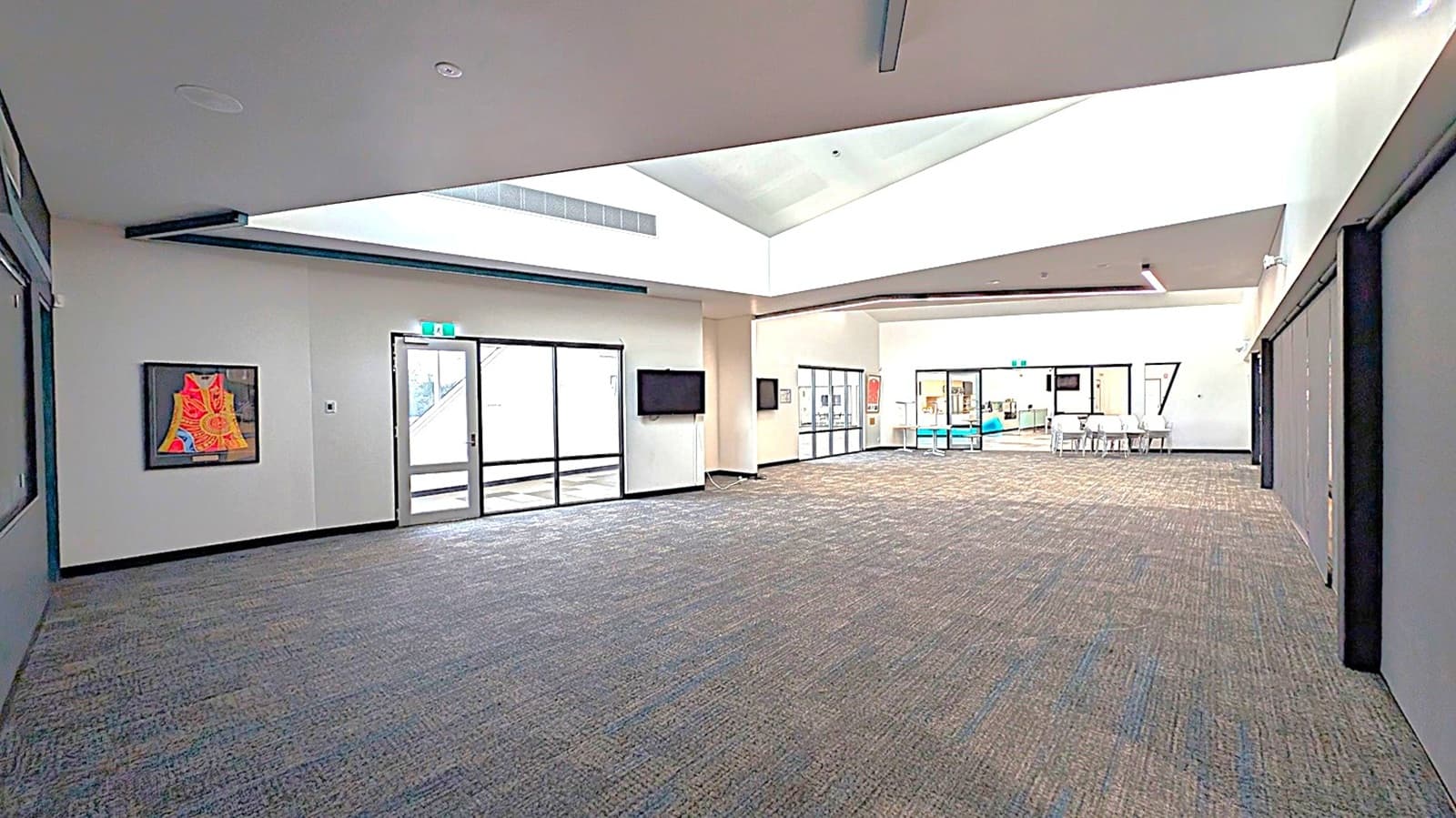 A spacious interior with grey carpeted flooring. On the left, there's a colourful framed artwork on the wall and a glass door leading outside. A wall-mounted TV is nearby. In the centre, a large open area with a high ceiling and skylight. On the right, glass doors open to a room with white chairs and a turquoise seating area. The ceiling features recessed lighting.