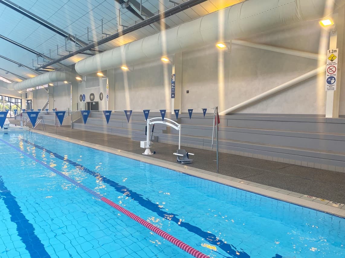 The pool features steps with handrails and colourful floating ropes. A hoist is positioned on the pool deck on the right. Blue triangle banners hang across the pool. Tiered seating is on the right. Adjacent to the pool are signages for accessible toilets. The ceiling features exposed metal beams with large aircon vents and light fixtures. Reminder posters are affixed to the wall.