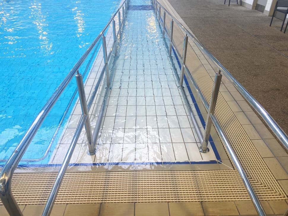 The pool features a ramp with handrails. The pool deck is lined with a pool overflow grid parallel to the ramp. Chairs are set against the wall to the right.
