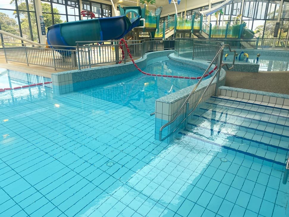An indoor swimming pool with clear blue water. A blue water slide with a yellow entrance is positioned to the left, partially visible. The pool has a raised area to the right with a round, wading pool section separated by a red rope divider. Stainless steel railings lead into the shallow end of the pool. Surrounding the pool are glass-paneled walls allowing a view of the trees outside. Safety posters are affixed to the wall.