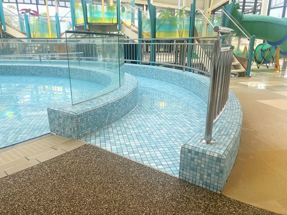 A swimming pool area with a mosaic-tiled pool with gentle curves shaping the pool edges and steps leading down into the water. To the right, there is a metal railing, presumably for safety and assistance. The floor around the pool is speckled with brown, beige, and white tones. In the background, parts of water slides in green and blue are visible, along with a transparent structure, likely a tube slide.