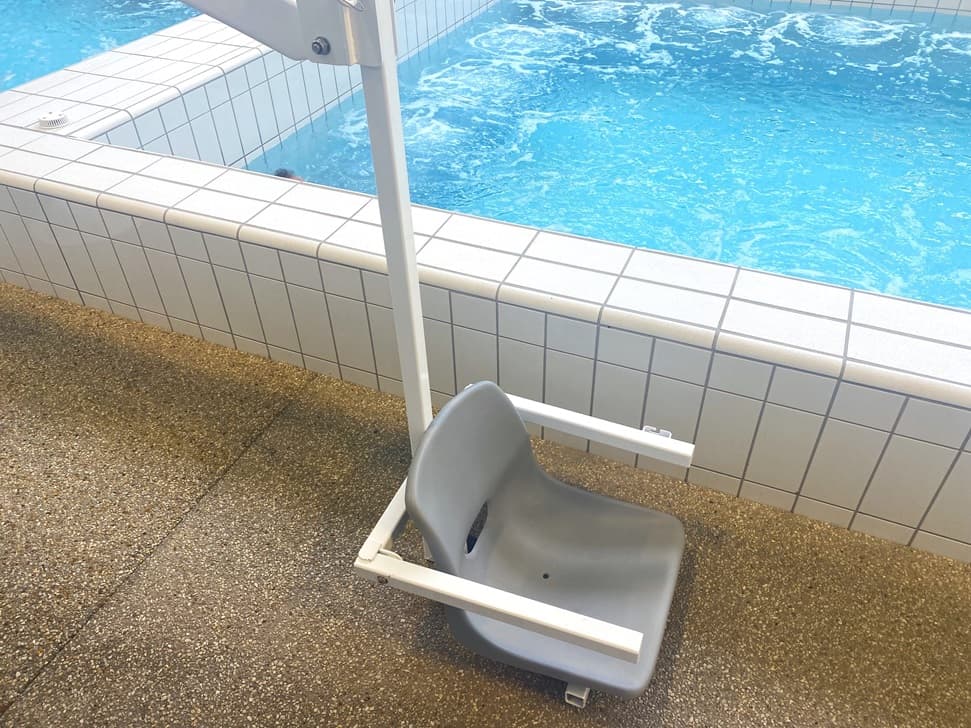 A hoist with a chair.Beside is a pool with white tiles coping. The floor around the area is speckled with brown, beige, and white tones.