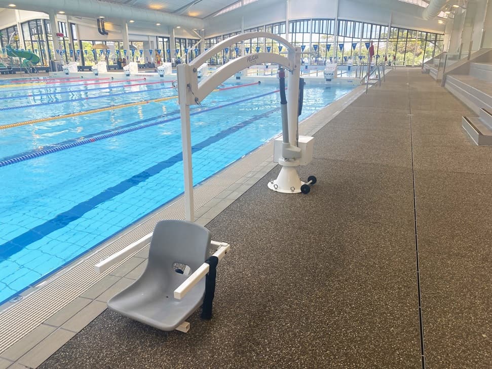 A Pelican hoist with a chair is positioned on the pool deck. Beside it, the lap pool features steps with handrails and colourful floating ropes. A string of blue triangle banners hangs across the pool. Tiered seating is on the right, with a view of the children's pool visible at the far end. Glass surrounds the space.