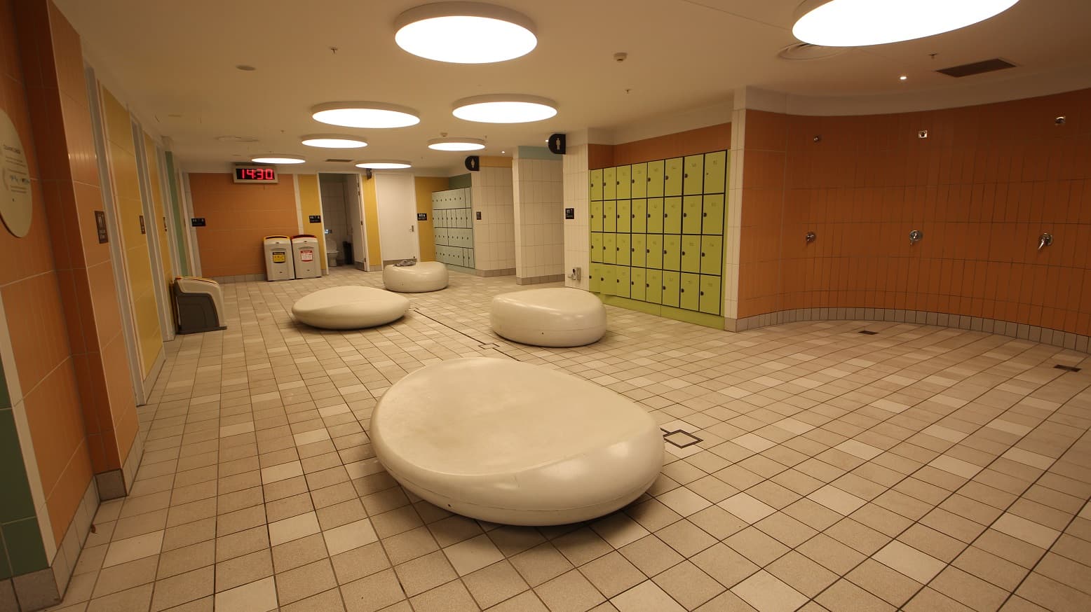 Leading to the Changing places facility at Yawa Aquatic Centre. In the open space are large-looking stones with slightly carved middle portions. The floor tiles are light brown with some random white sections. The shower area has an orange tile wall. This facility is illuminated by large circular lights that are elevated from the ceiling. There is a large digital clock mounted on the orange tiled wall at the back and two locker cabinets next to the entry corridors.