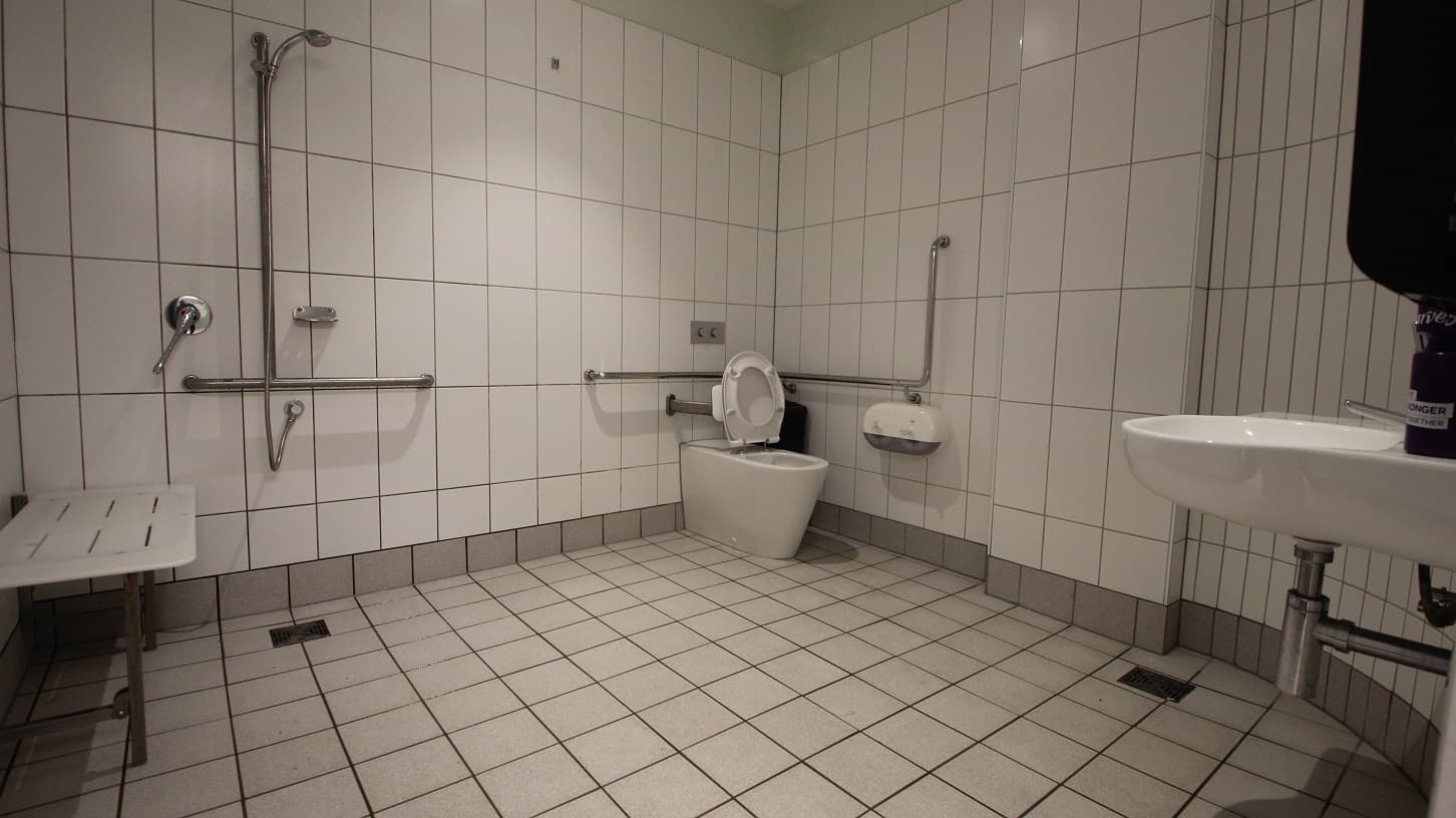 Near the shower area to the left is a small white platform for sitting. Opposite it is the accessible toilet seat with a white cushion to support the back of the person, a grab bar, and a white toilet paper holder. There is also a wash basin with liquid hand soap. The bathroom has a white tiled wall with a lime green portion before the ceiling and a grey tiled floor.