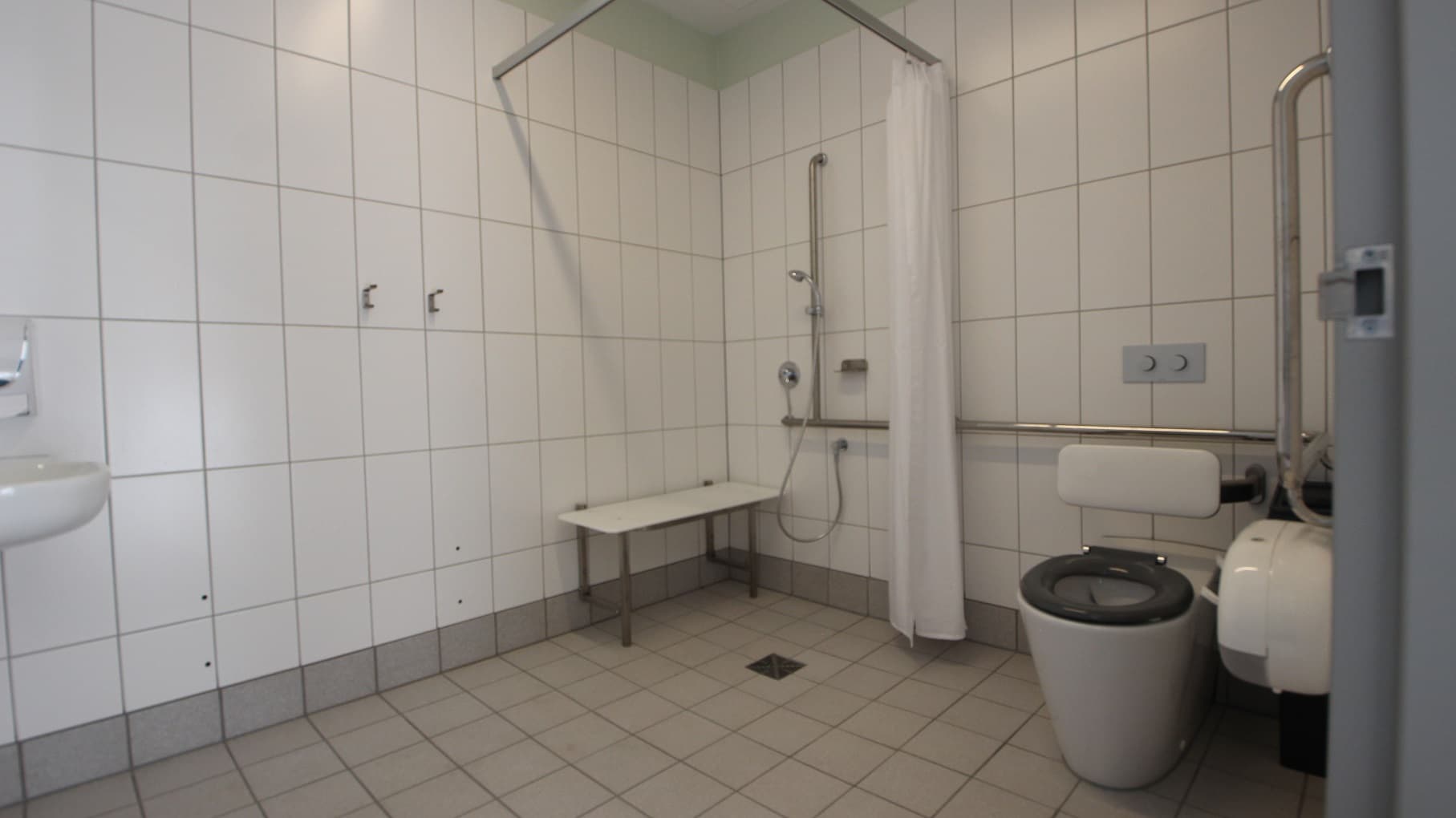 Another view inside one of the accessible toilets at Yawa Aquatic Centre. The shower area with a white platform for sitting has a white privacy curtain that can cover the entire shower section. The toilet seat has a grey cover with a white back cushion. There is also a white toilet paper holder mounted near the toilet.
