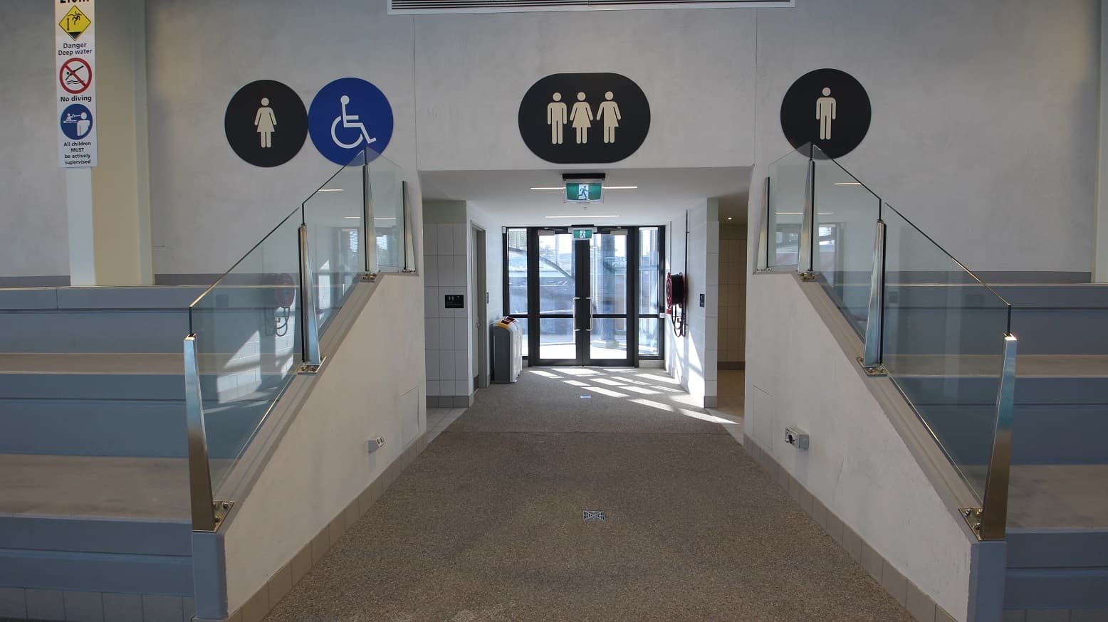 Pathway in between the grey seating stadium with metal and glass handrails at Yawa Aquatic Centre. On the white structural post on the wall of the seating stadium is a white rectangular sign board for “danger deep water”, “no diving”, and “all children must be actively supervised”. There are large circular signboards on the grey wall of the seating stadium. Near the glass doors are two rubbish bins.