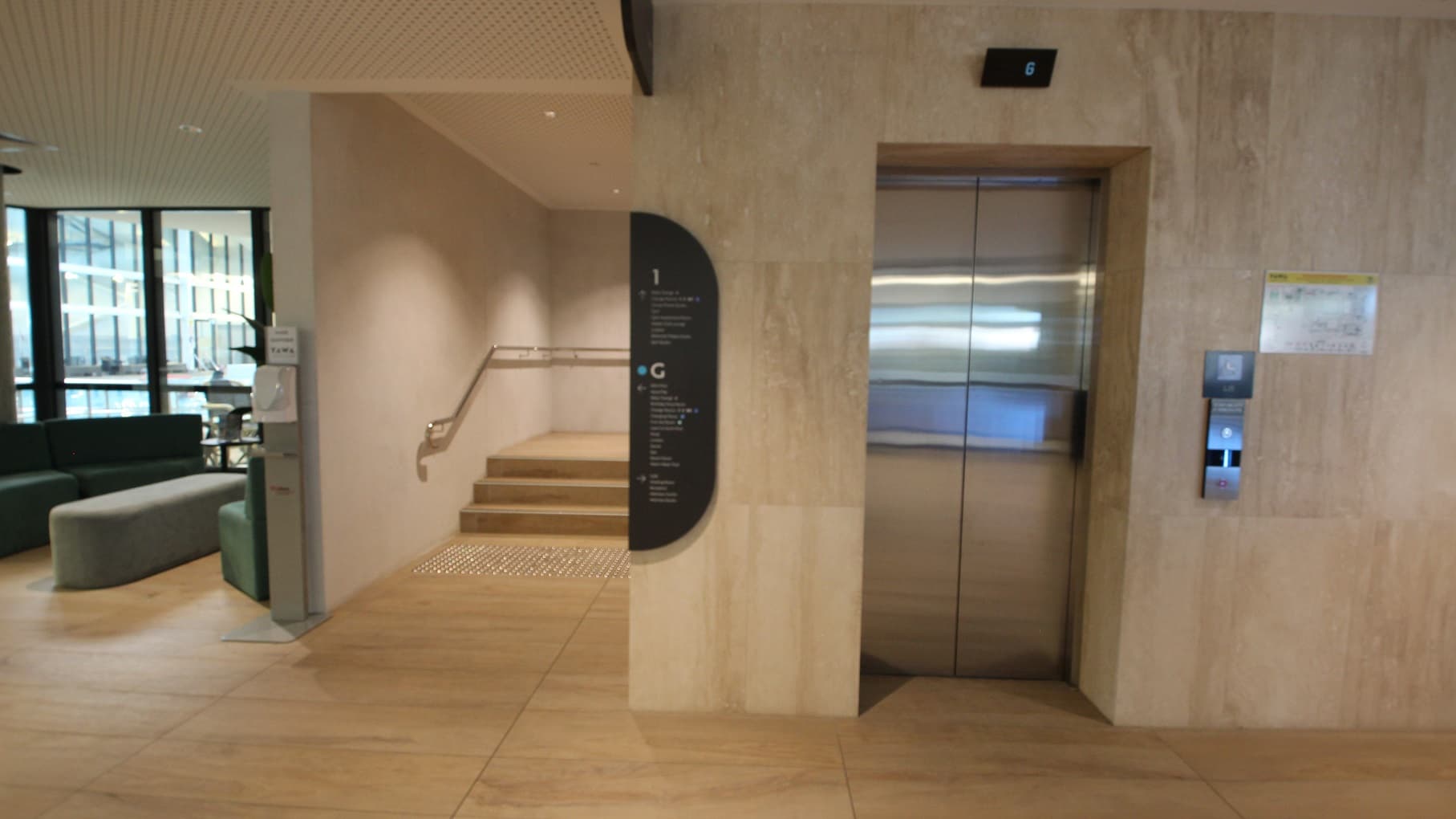 Lift access to the Health Club on Level 1 at Yawa Aquatic Centre. On the left is a seating area with sofa chairs with a view of the aquatic facilities. Next to the lift is a staircase with metal handrails placed on the wall. There is a black sign board that contains the different facilities on each floor to guide guests. A black notification screen on top of the lift can be seen for lift tracking.