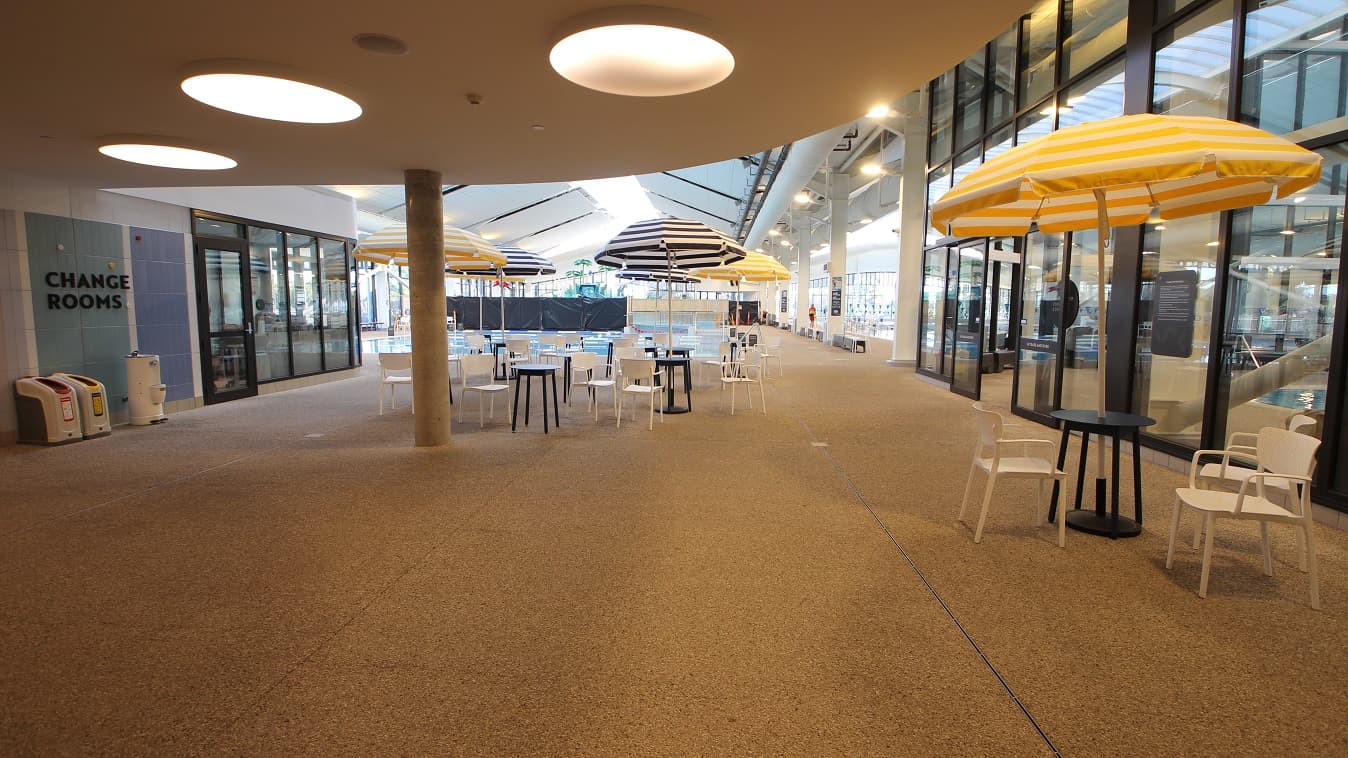A series of blue tables and white chairs with yellow or blue umbrellas create a light summery feel inside the aquatic facility of Yawa Aquatic Centre. On the left side are the Change rooms, which can be identified by the varied colors of the tile wall. This section of the facility has a granite floor with a curvy ceiling with large circular fluorescent lights. There is a pool at the back with metal benches on the side. A black wall next to the pool is noticeable from this position.