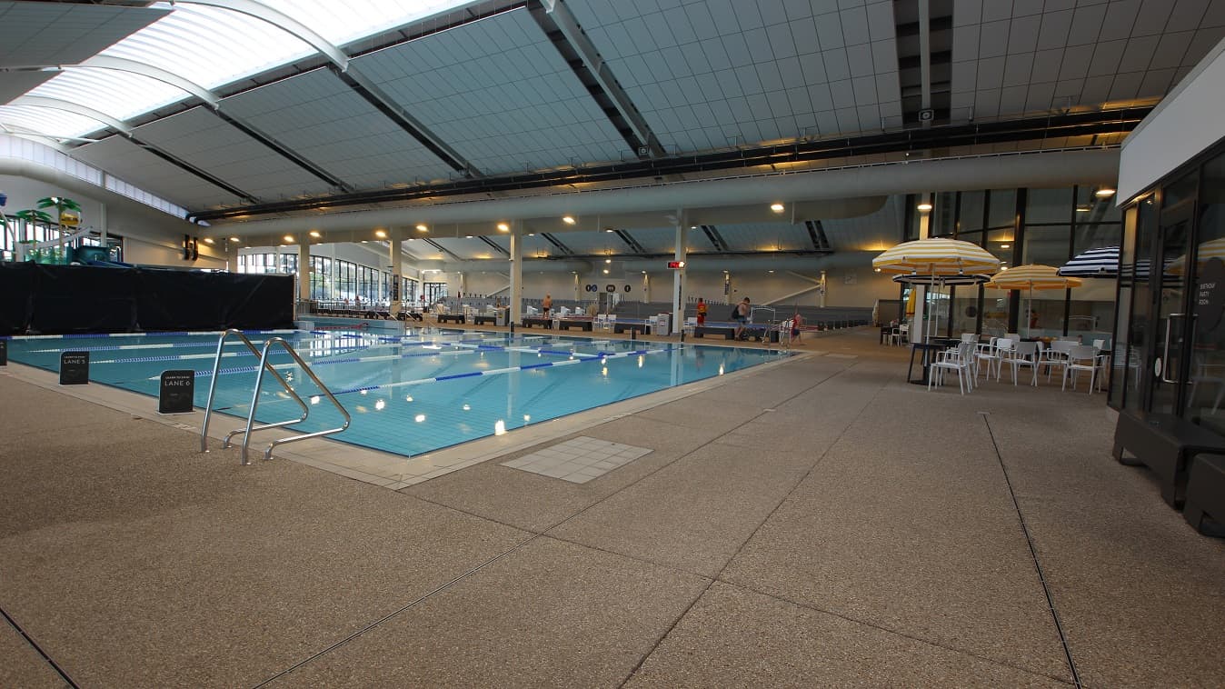 Side view of the Learn to Swim pool at Yawa Aquatic Centre. On the left is the pool ladder marked by a curvy metal structure at the edge of the pool. The facility has dome-like roofing with natural light filtering through the focal point of the roof. The pool has a staircase with metal handrails. Each lane has a black signboard at the starting point and is separated by white and blue floating lane ropes. A black wall next to the pool separates the Kid's Waterslides and Splash Zone.