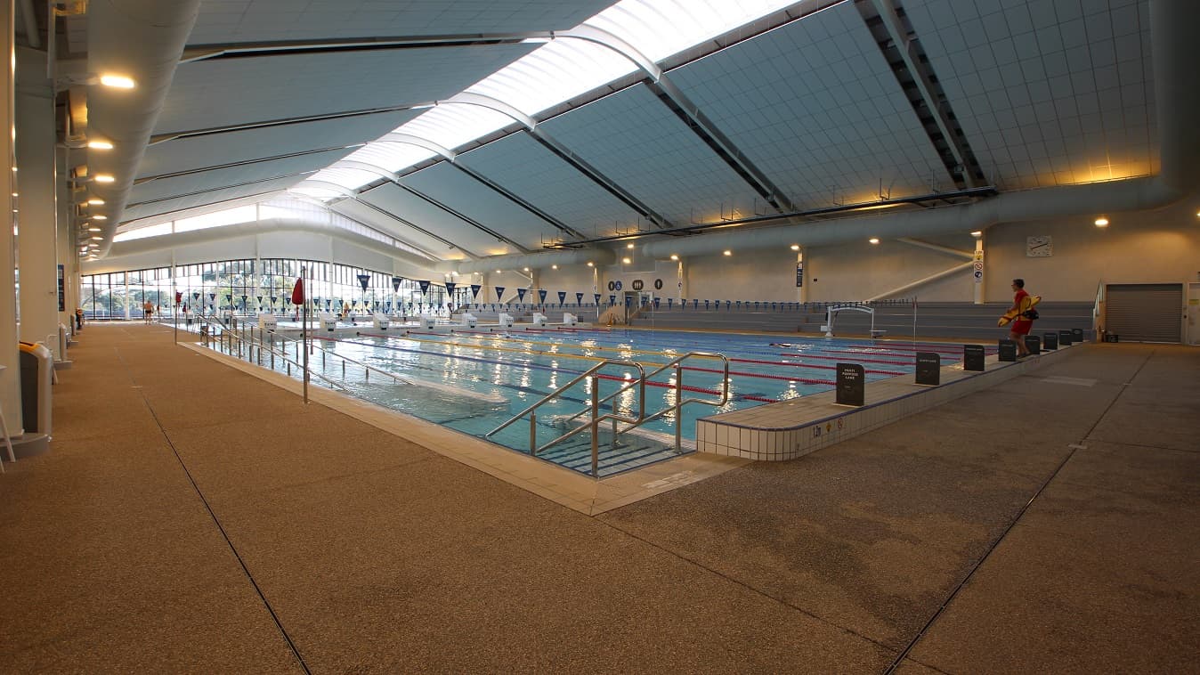 The 8-lane 50m pool at Yawa Aquatic Centre. There is a railed entrance ramp and railed staircase directly opposite each other on the left side of the pool. The lanes are separated by yellow, blue, and red floating lane ropes. Near the staircase is an elevated section with black signboards. On the opposite end are white diving boards while near the grey seating stadium is a white pool hoist. Intersecting horizontally to the pool is a string of blue flags tied to opposite poles of the pool.