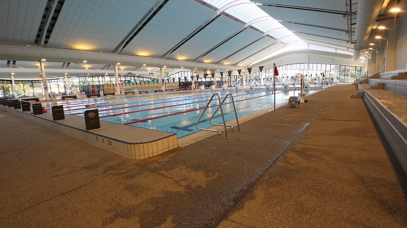 View of the 8-lane 50-meter pool from the grey seating stadium. There is a pool ladder marked by a curvy metal structure. The white hoist has a grey chair to help move persons with mobility disabilities. The entire aquatic facility has glass walls that enable natural light to filter through the inside and at the same time provide a great view of nature. A lifeguard in a yellow and orange uniform is actively patrolling the pool area.