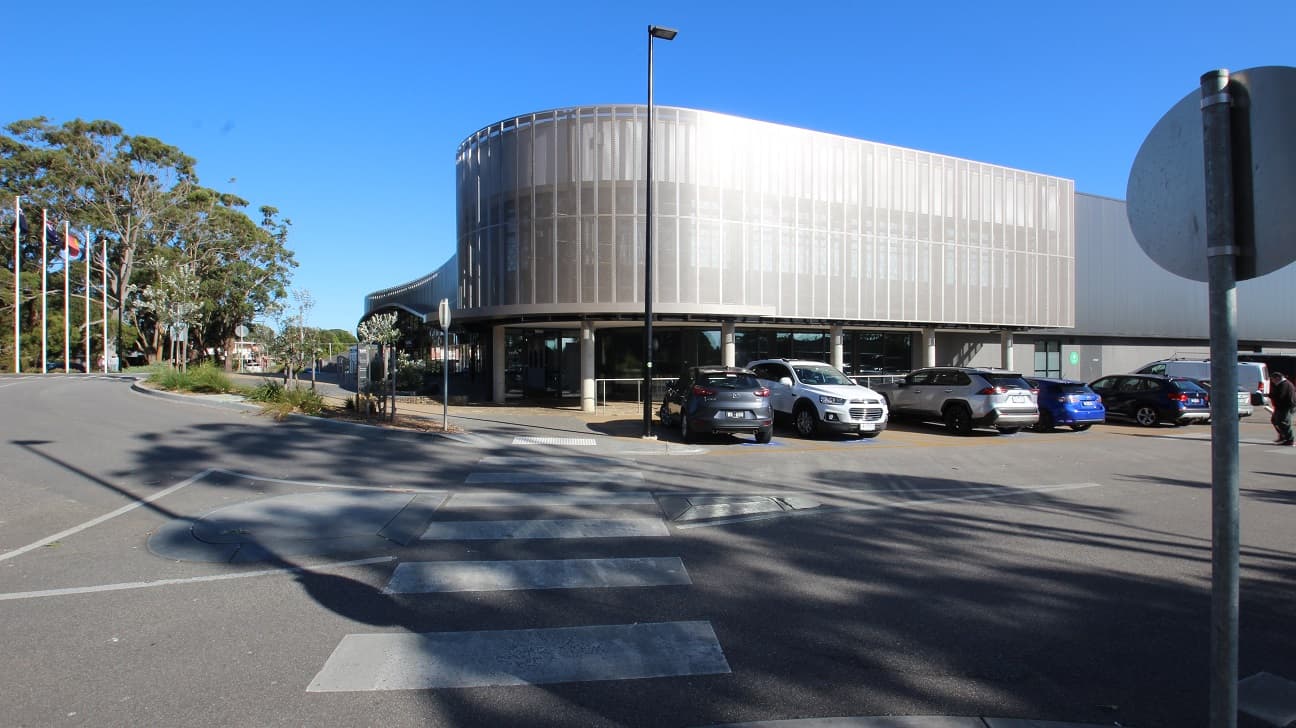 Zebra crossing at the accessible car parking space of Yawa Aquatic Centre. At each end of the crosswalk is a metal section that is part of the triangular line of the driveway. There are several cars parked in the parking space. From this position, seen on the opposite road are the tall trees with canopy cover. Small plots for plants surrounding the facility are placed for landscaping.