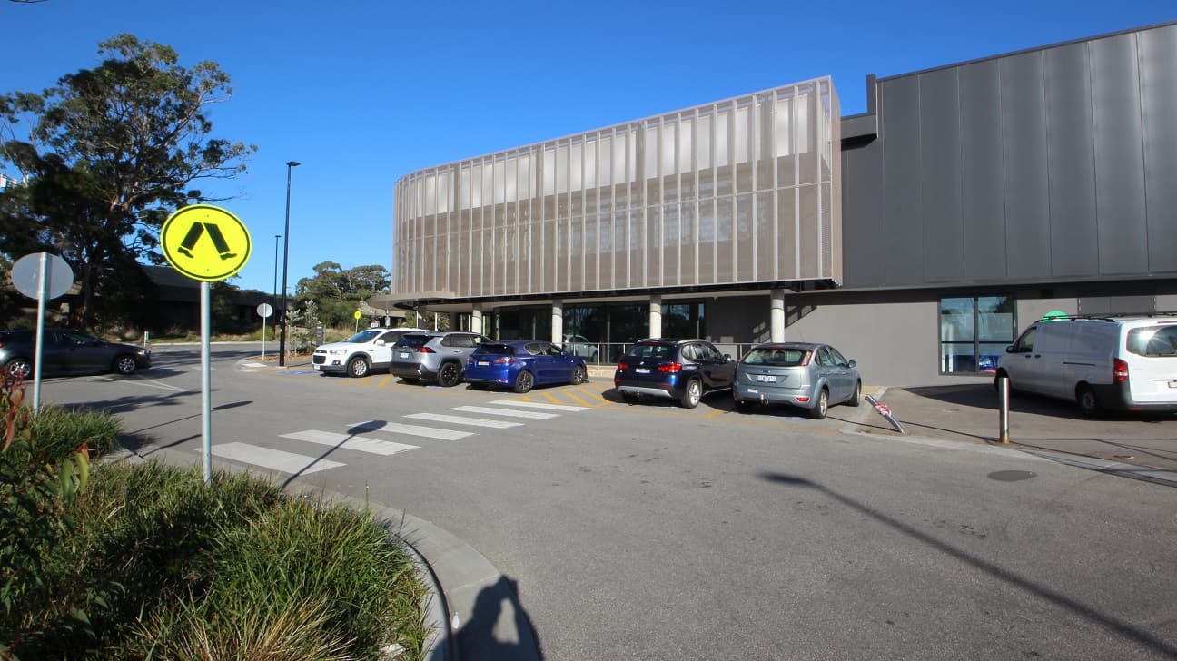 Another zebra crossing within the car parking space of Yawa Aquatic Centre. Placed before each crossing are two yellow pedestrian crossing signs facing in opposite side directions. The planter box is filled with healthy shrubs and bushes. The accessible car parking spaces can be distinguished by the yellow lines and is near the entrance of the facility. The soft curvy pattern covering the wall ends here, and the walls can be seen painted in grey.